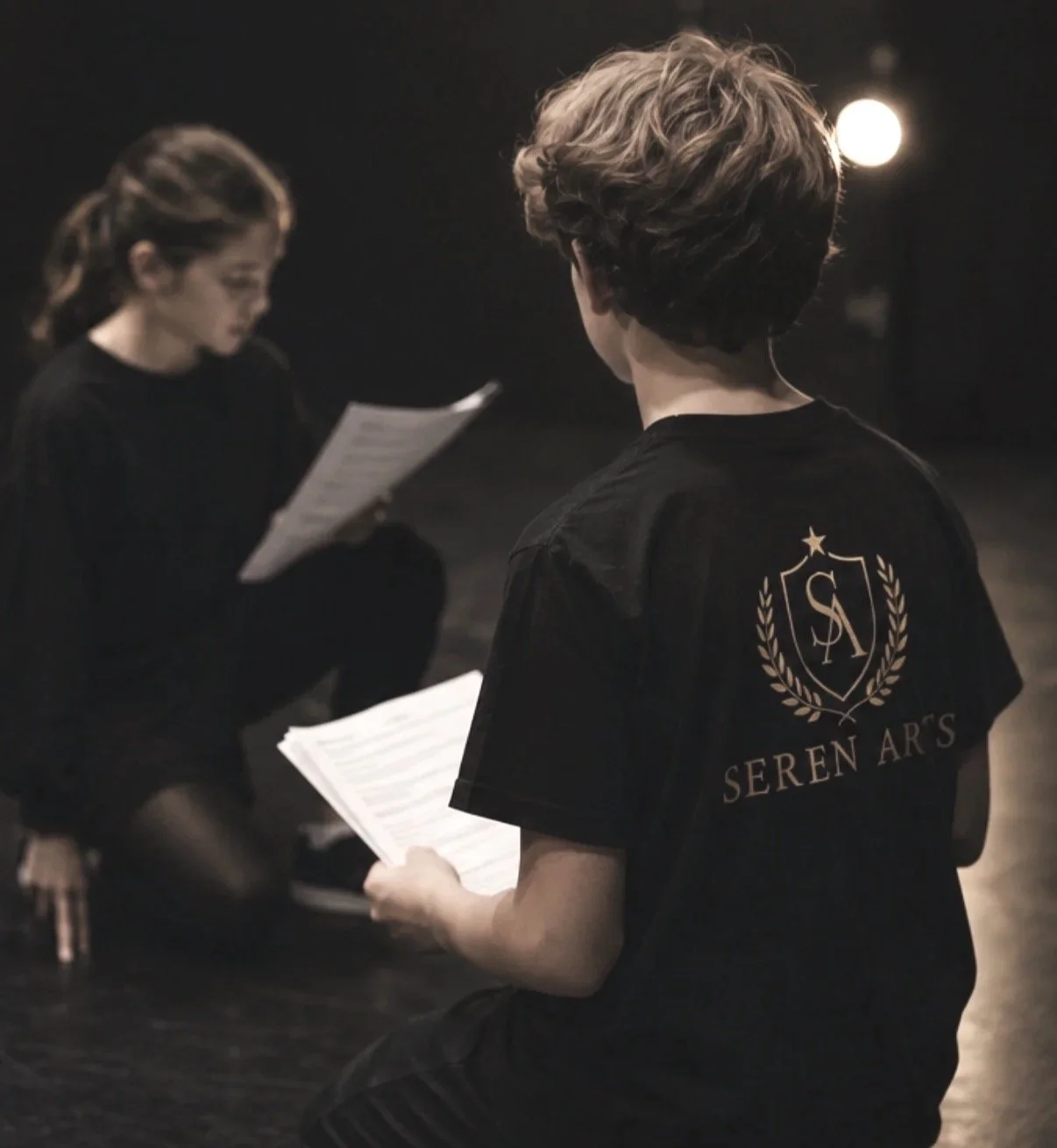 Two young children, a boy and a girl, are sitting on the floor in a dark room, holding and reading sheets of paper. The boy's shirt has a crest with initials and the words 'Seren Arts' on the back.