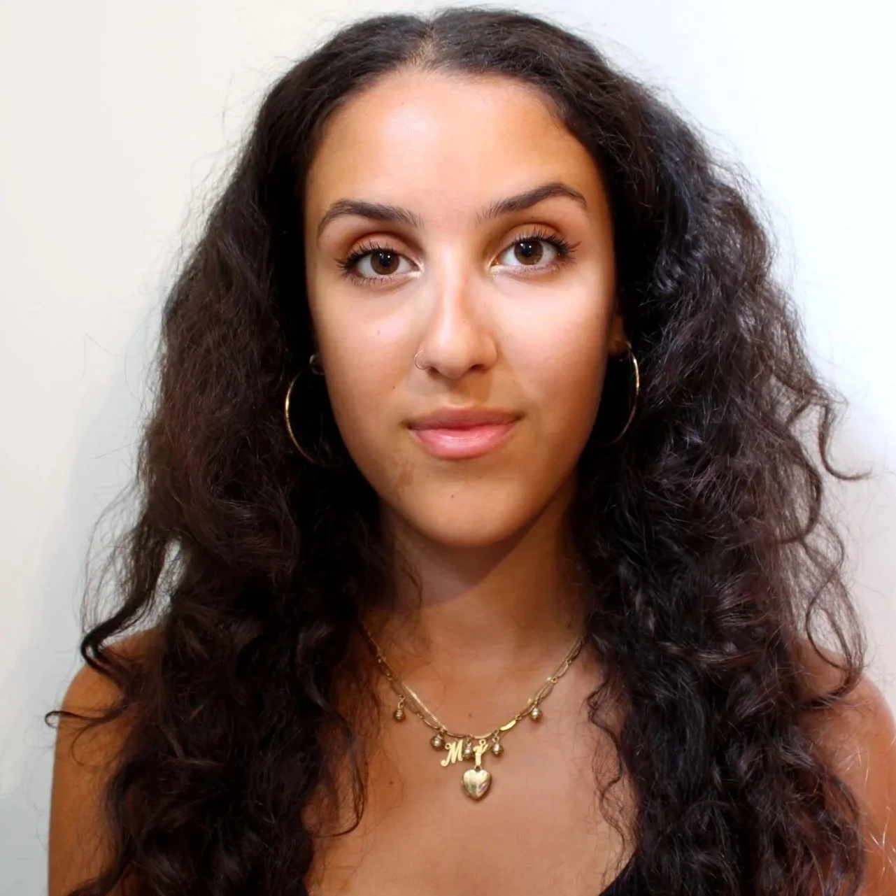 A young woman with long curly dark hair, wearing gold hoop earrings and layered gold necklaces, poses against a plain white background.
