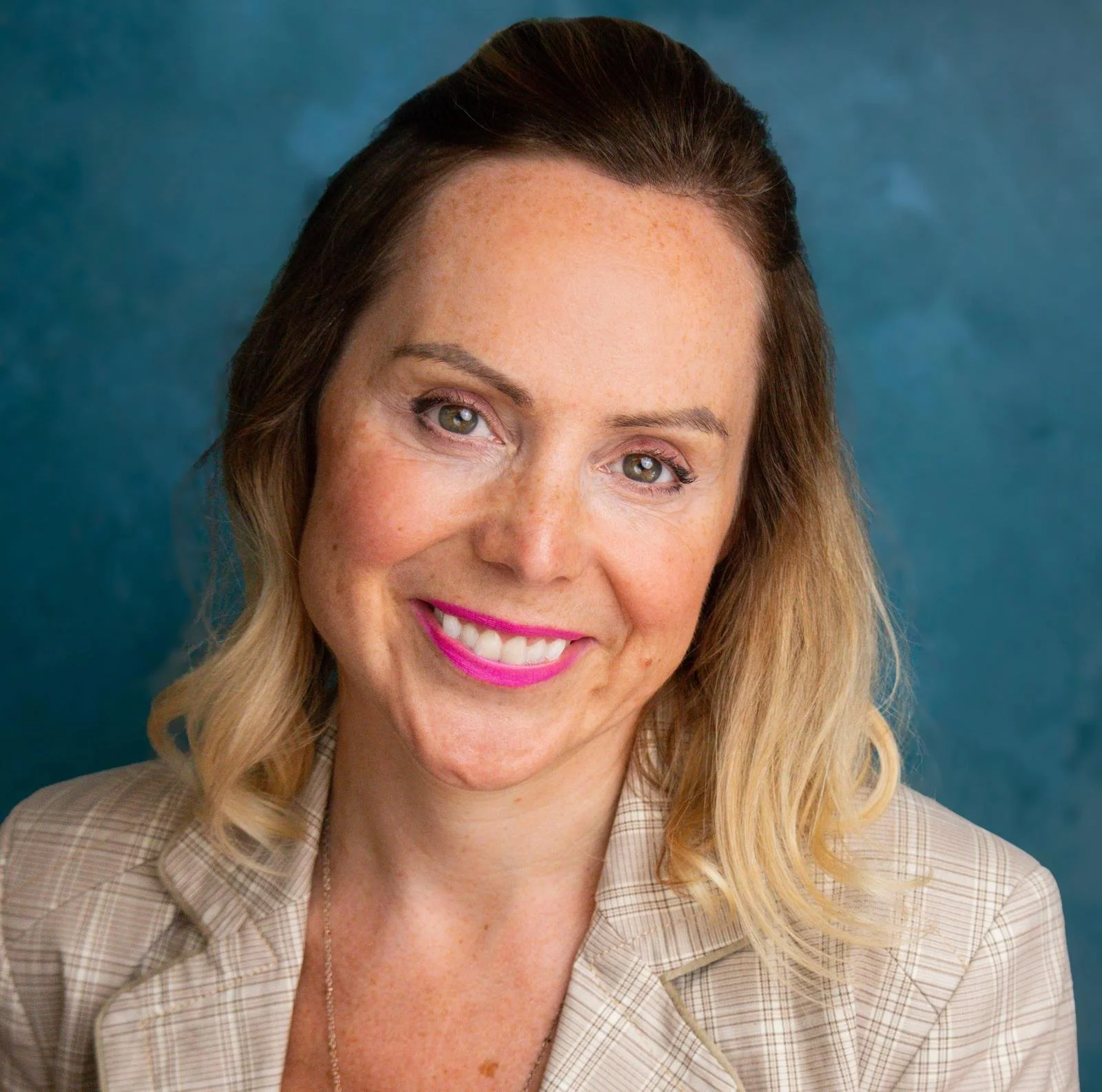 Close-up of a woman with blonde hair, wearing a checkered blazer, smiling with bright pink lipstick, against a blue background.