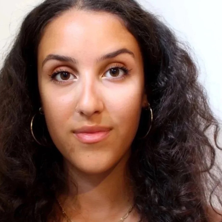 Close-up photo of a young woman with curly dark hair, wearing gold hoop earrings, a nose ring, and layered gold necklaces, looking at the camera with a neutral background.