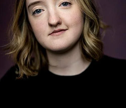 A young woman with shoulder-length blonde hair, blue eyes, and a neutral expression, wearing a black top against a dark purple background.