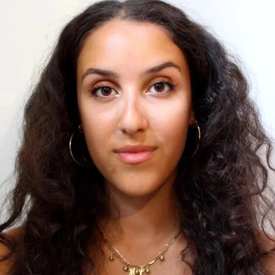 Close-up photo of a young woman with curly dark hair, wearing gold hoop earrings, a nose ring, and layered gold necklaces, looking at the camera with a neutral background.