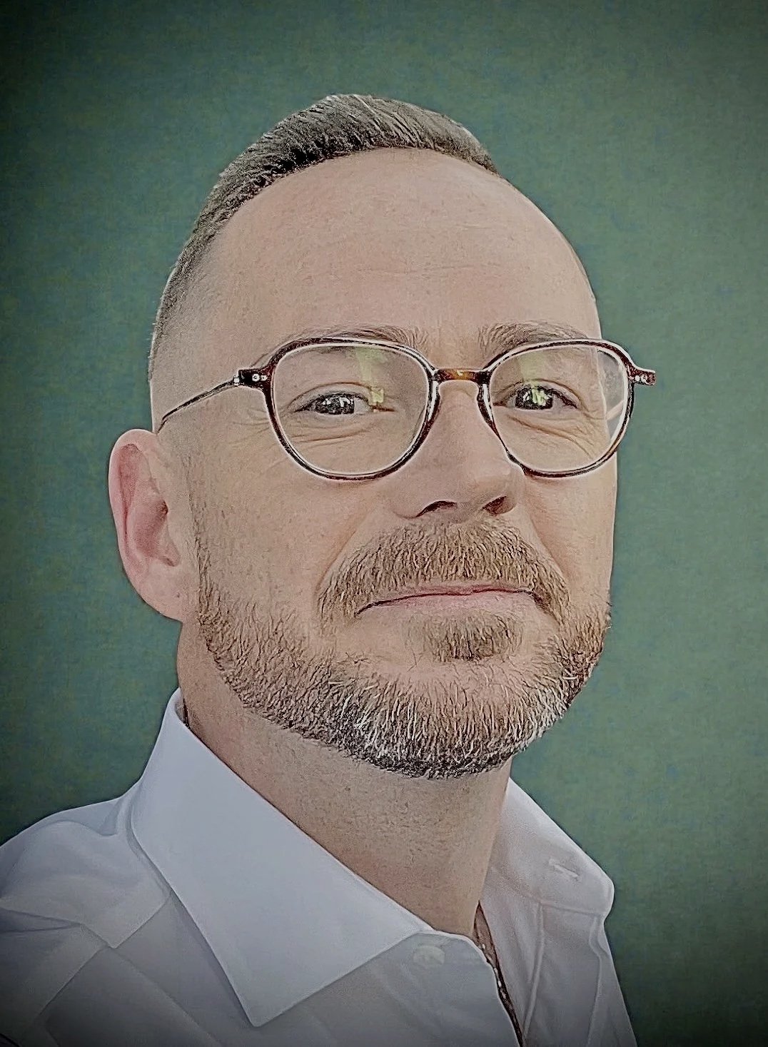 A man with short dark hair, glasses, a beard, and a mustache, wearing a white shirt, posing against a green background.