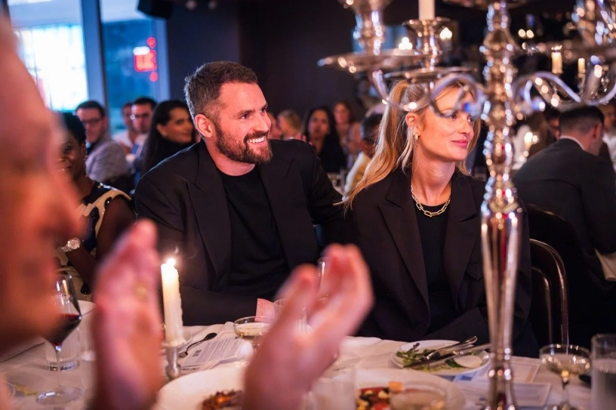 People attending a formal dinner event, seated at a table with a candelabra centerpiece, in a dimly lit room with large windows, with a man and woman in focus.