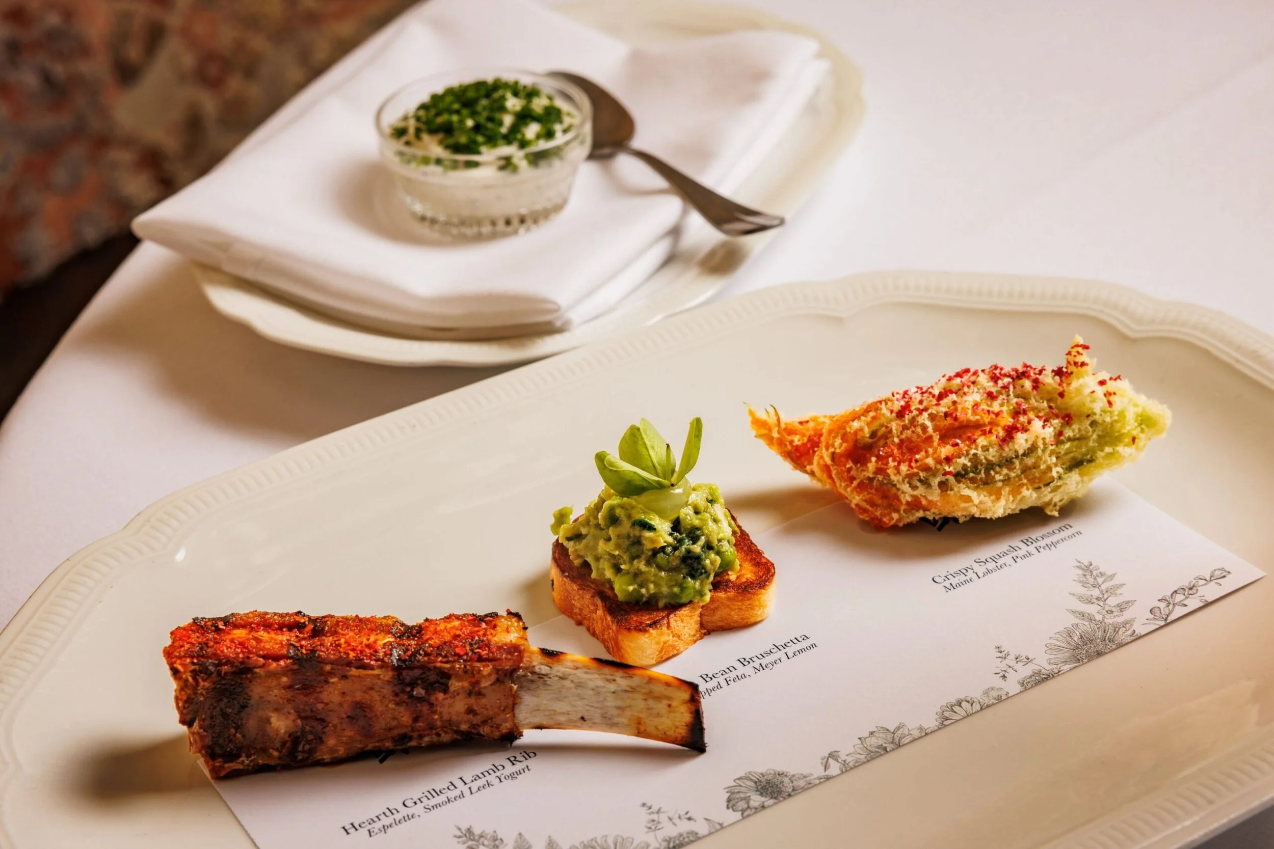A white plate with three appetizers including guacamole on bread, a slice of lasagna, and a slice of salmon loaf. In the background, there's a small bowl of what appears to be sour cream with chopped herbs, and a fork resting on a white napkin.