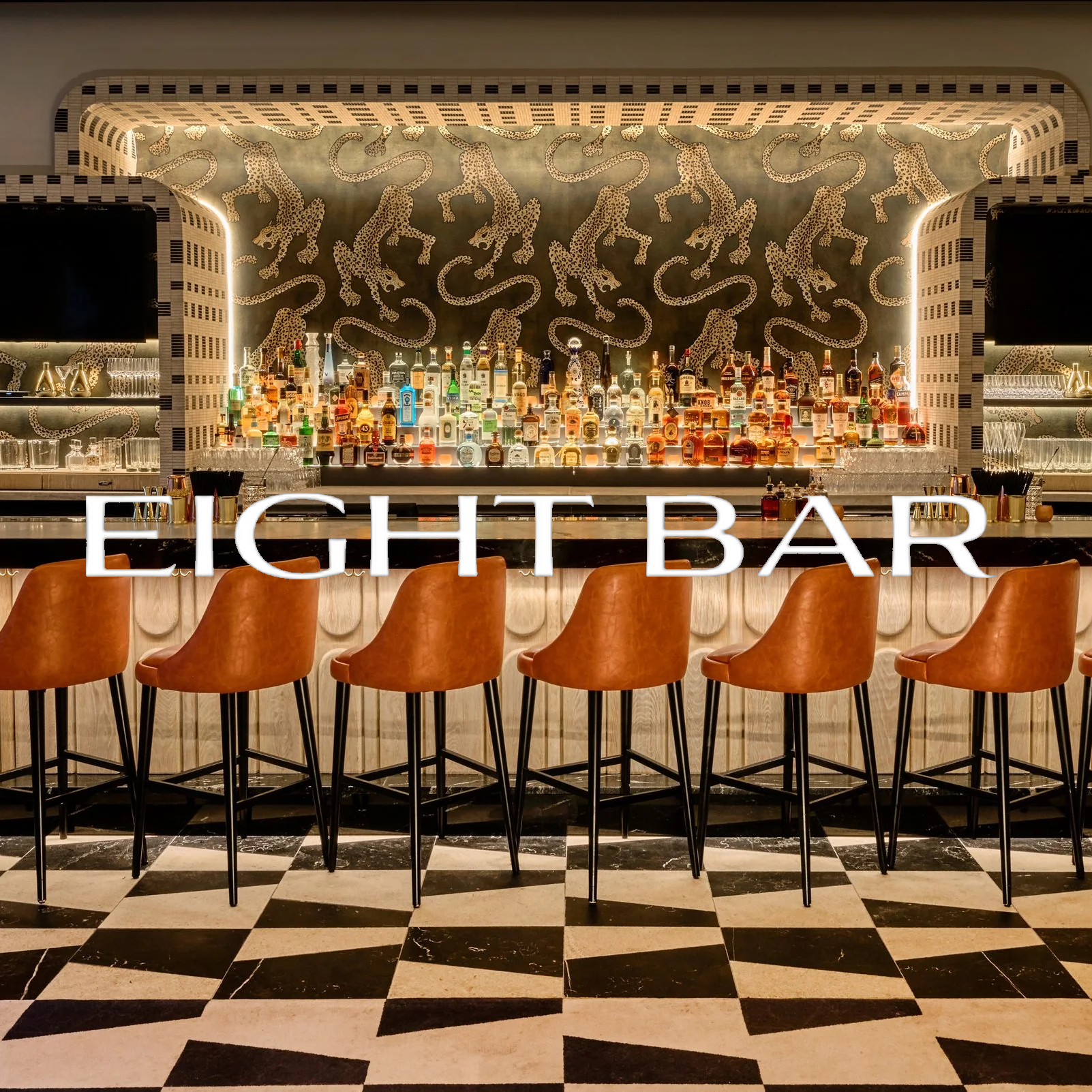 A stylish bar with a black and white checkered floor, a backlit display of various liquor bottles, and a wall with a pattern of leaping cheetahs. There are eight orange bar stools in front of the counter, with glasses and bar tools on the counter.