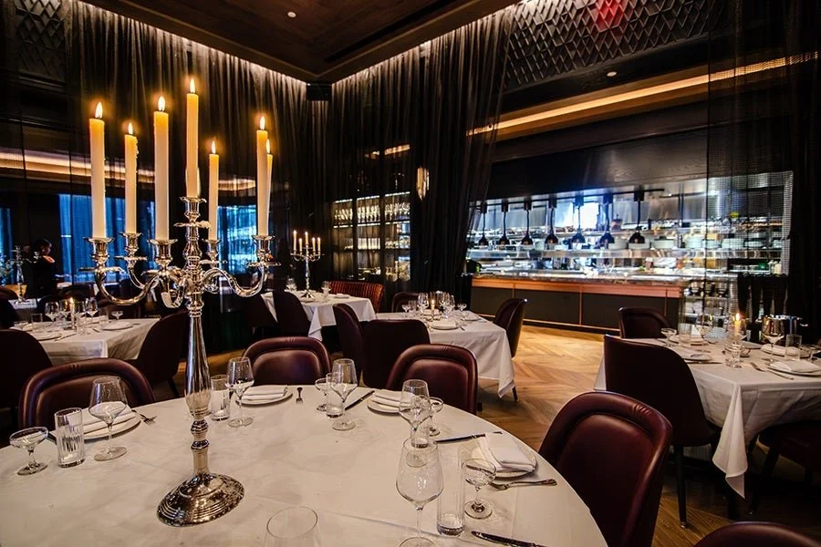 Elegant restaurant with round tables covered in white tablecloths, set with glasses, silverware, and napkins; a large silver candelabrum with lit candles on one table; dark chairs around the tables; a glass display case in the background with food and drinks; dark curtains and modern lighting.