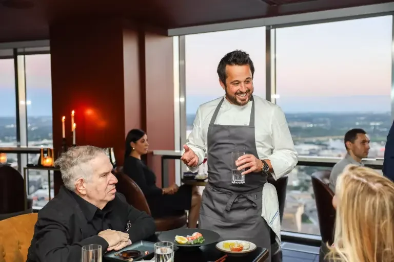 A smiling waiter with a glass of water attending to guests at a restaurant with a city view in the background.