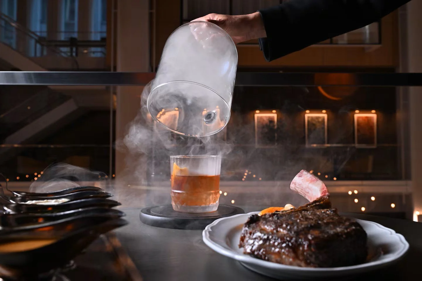 A chef pours a smoky drink into a glass on a restaurant table, with a cooked piece of meat on a plate in the foreground.