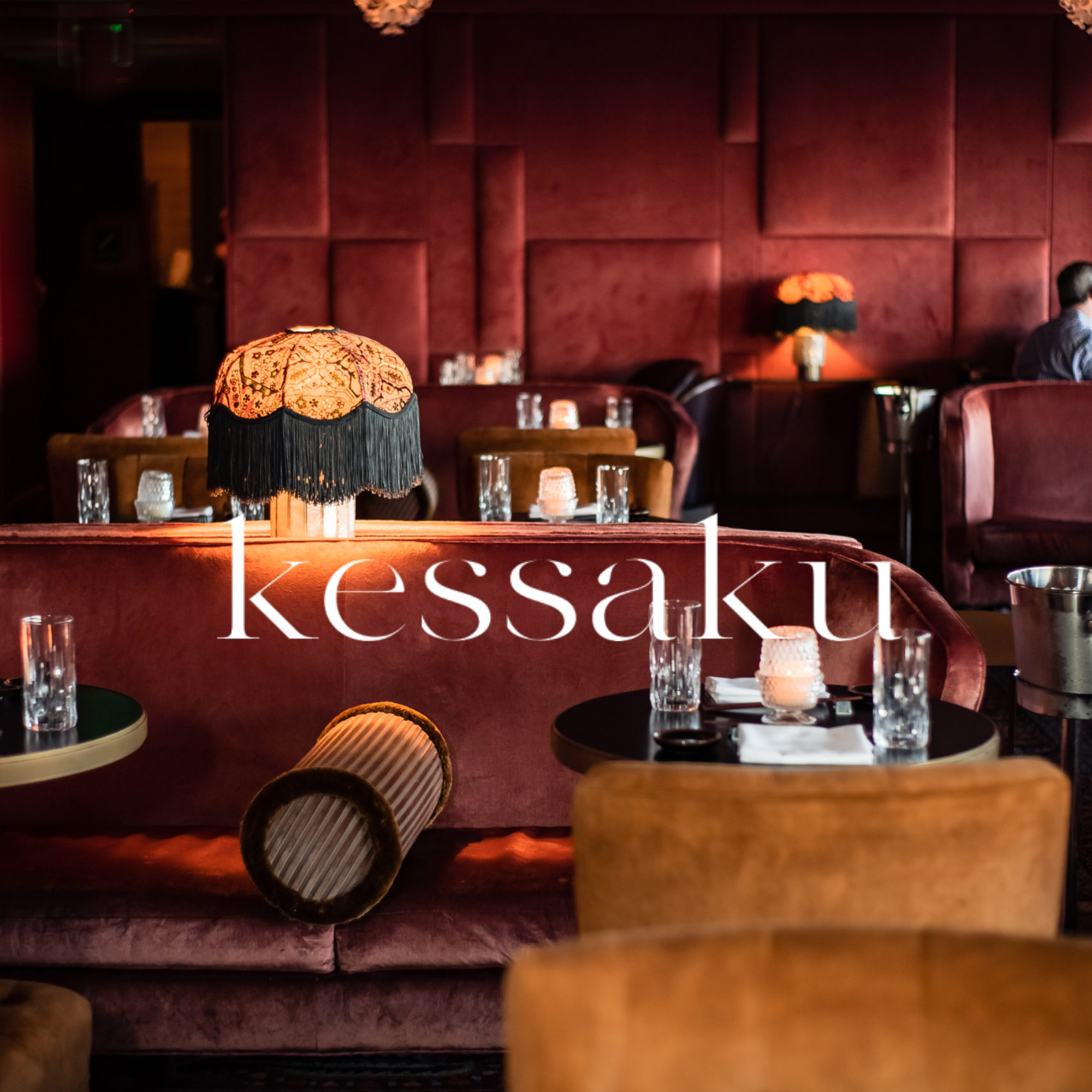 A warmly lit dining area with dark red upholstered walls and a table set with glasses and candles. There are decorative lamps with fringed shades, and a person with short hair sitting at a table to the right. The name 'Kessaku' is partially visible at the bottom of the image.