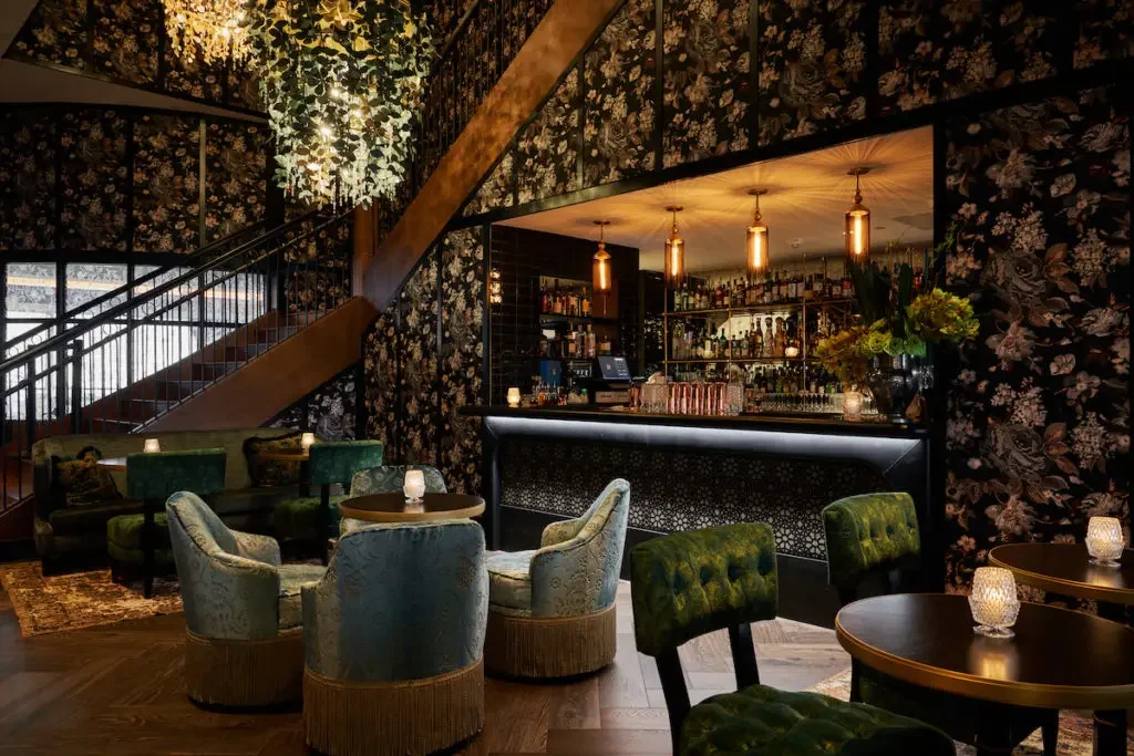 Interior of a stylish bar with floral wallpaper, plush chairs, a bar counter with bottles, warm lighting, and a staircase with greenery overhead.