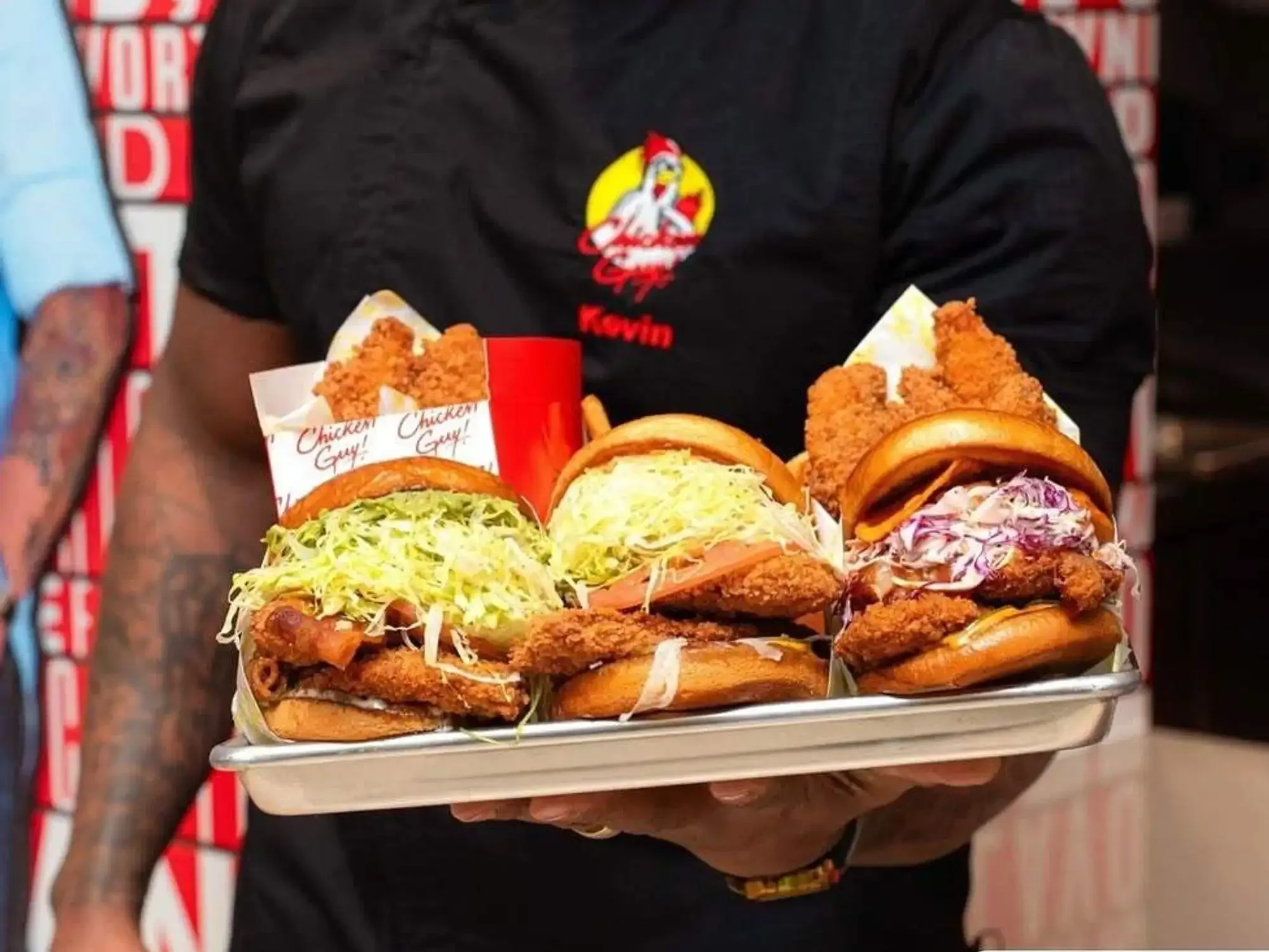 A person holding a tray with assorted fried chicken sandwiches topped with lettuce, coleslaw, and onions. The person is wearing a black shirt with a logo and the name 'Kevin' embroidered on it, and tattoos are visible on their arm.