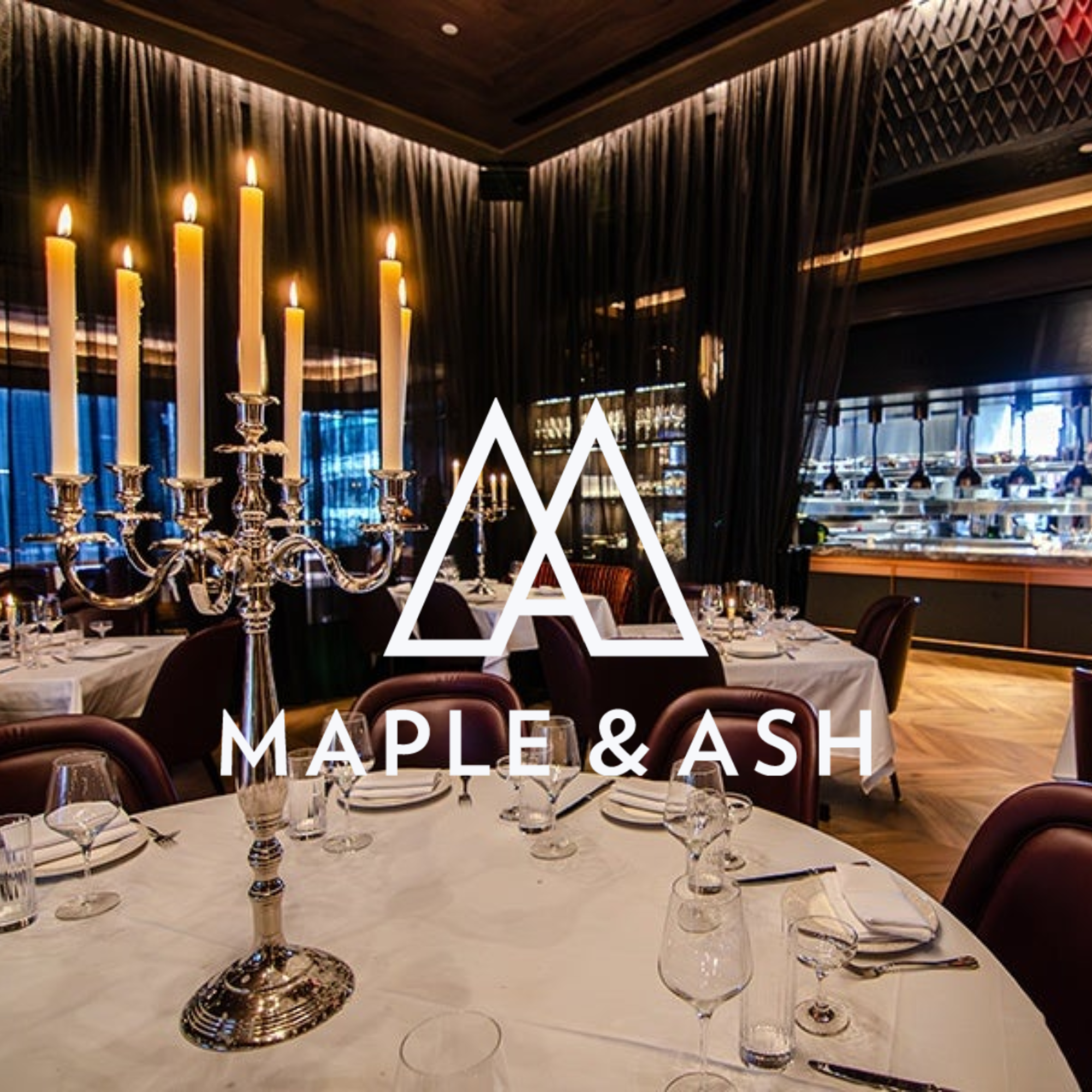 Elegant dining room with long tables set with white tablecloths, wine glasses, and silverware, featuring a large silver candelabrum with lit candles, and a background of dark wood ceiling and walls with modern lighting, large windows with blue curtains.