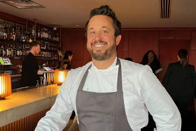 Smiling male chef in white uniform and gray apron in a restaurant bar area with bartenders and staff in the background.