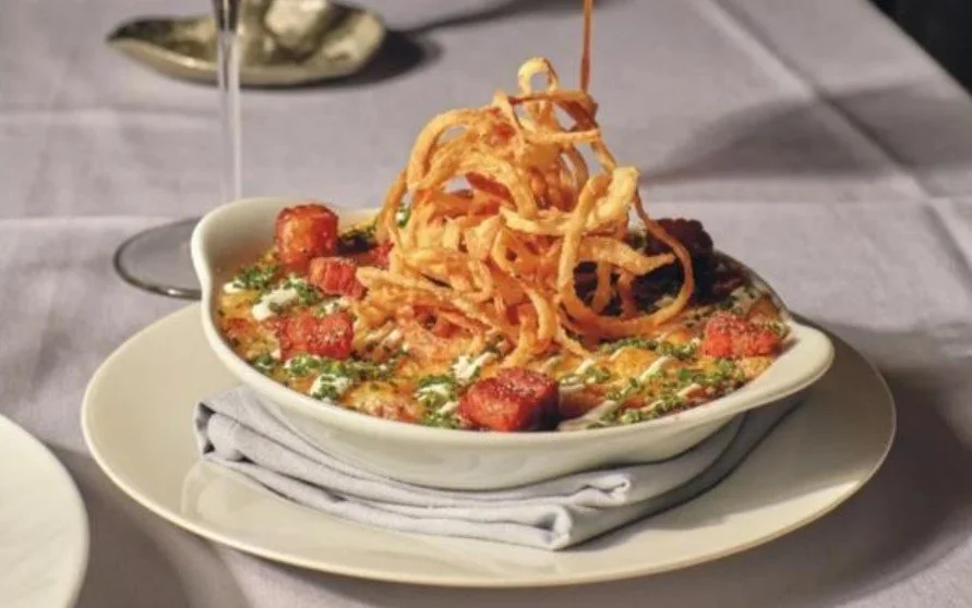 A bowl of loaded soup topped with crispy fried onion strings, with visible chunks of sausage and garnished herbs, served on a white plate with a napkin underneath.