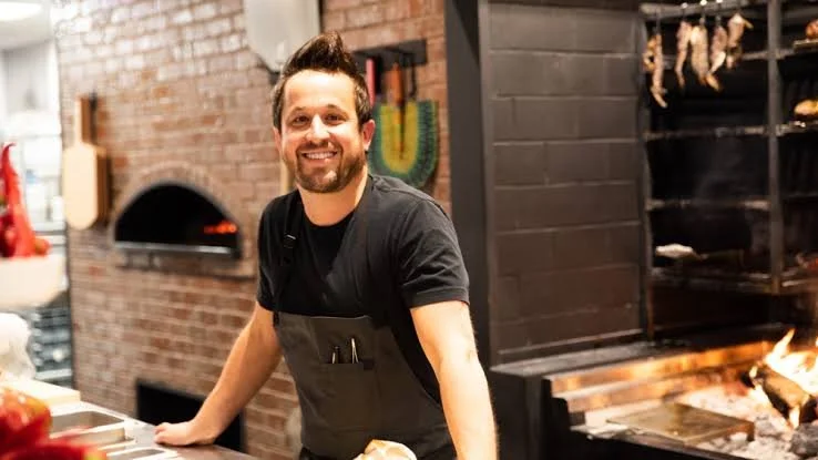 A smiling man working in a pizza restaurant with a brick oven in the background.