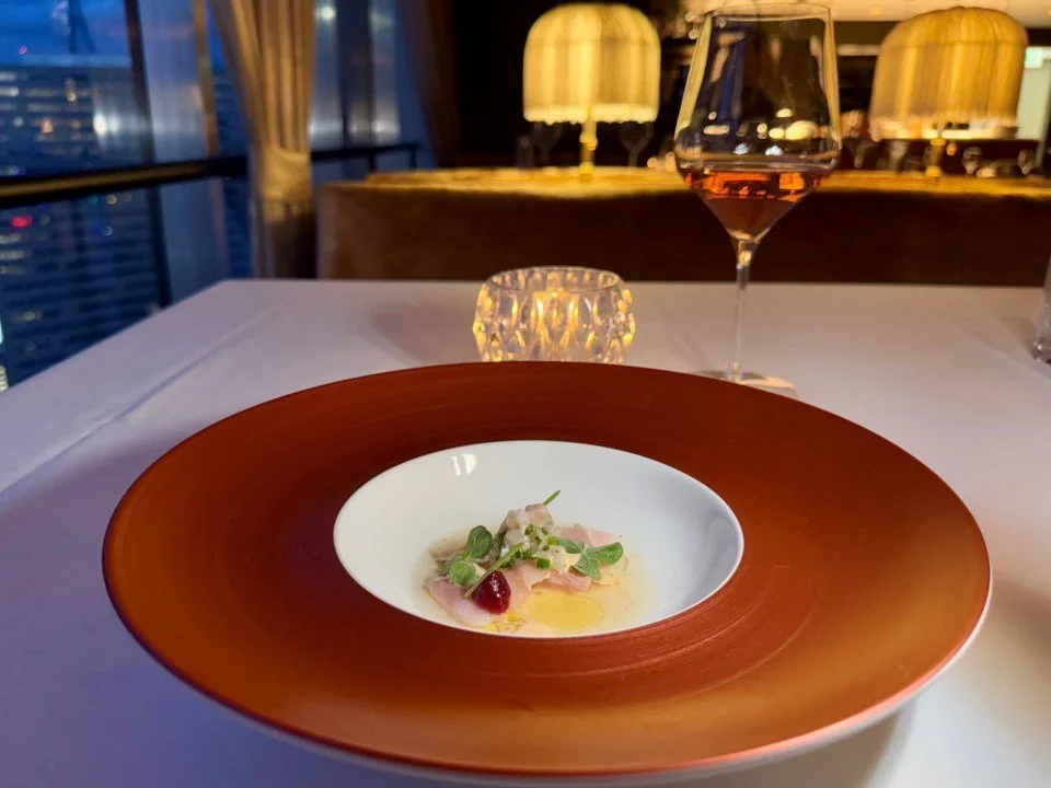 Fine dining table setup with a small gourmet dish, glass of rosé wine, a candle, and elegant lamps in the background.
