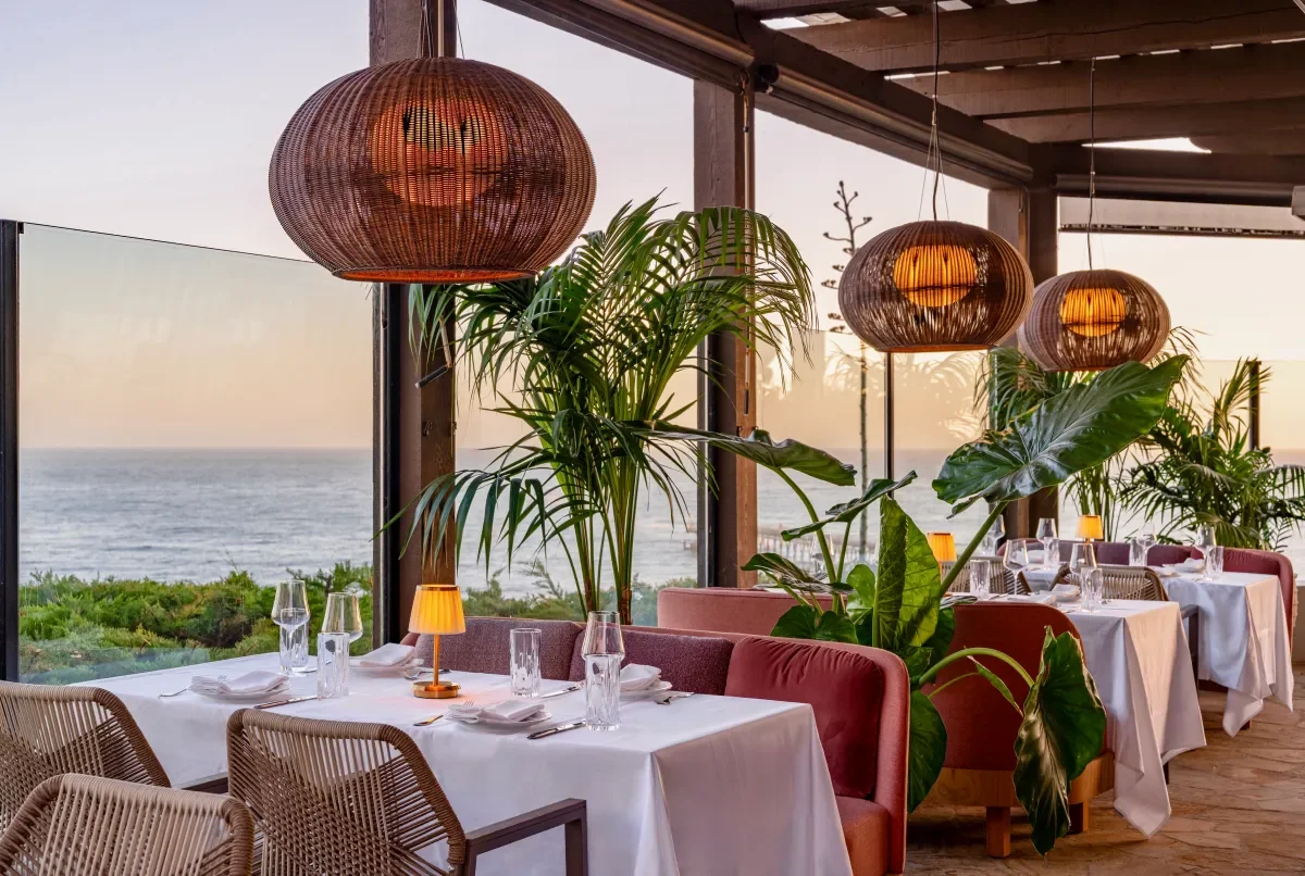 Elegant restaurant with white tablecloths, glassware, and napkins, decorated with green plants and warm pendant lights, overlooking the ocean at sunset.