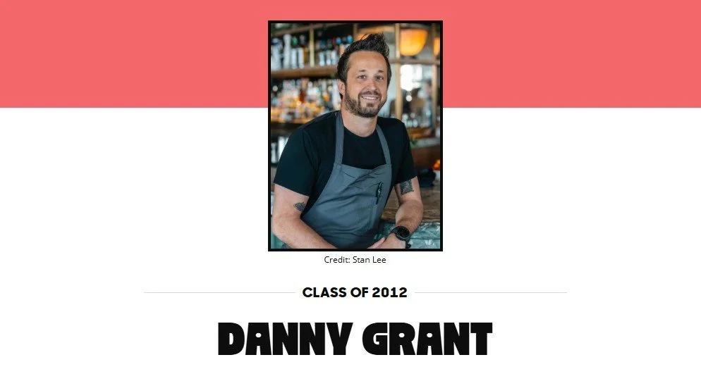 Photograph of a man with a beard and short dark hair, wearing a black T-shirt, gray apron, and a watch, standing behind a bar or counter in a bar or restaurant. The background shows shelves with bottles and glasses. Credit to Stan Lee. Text indicates he is from the class of 2012, and his name is Danny Grant.