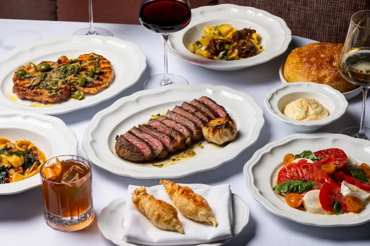 A table with various Italian dishes including a sliced medium-rare steak, spaghetti with meat sauce, tomato and mozzarella salad, ravioli, fried chicken tenders, a bread roll, and glasses of red wine and an aperitif.