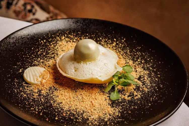 Dessert plate with a baked item topped with a golden sphere, garnished with green herbs, with crumbs scattered around on a black plate.