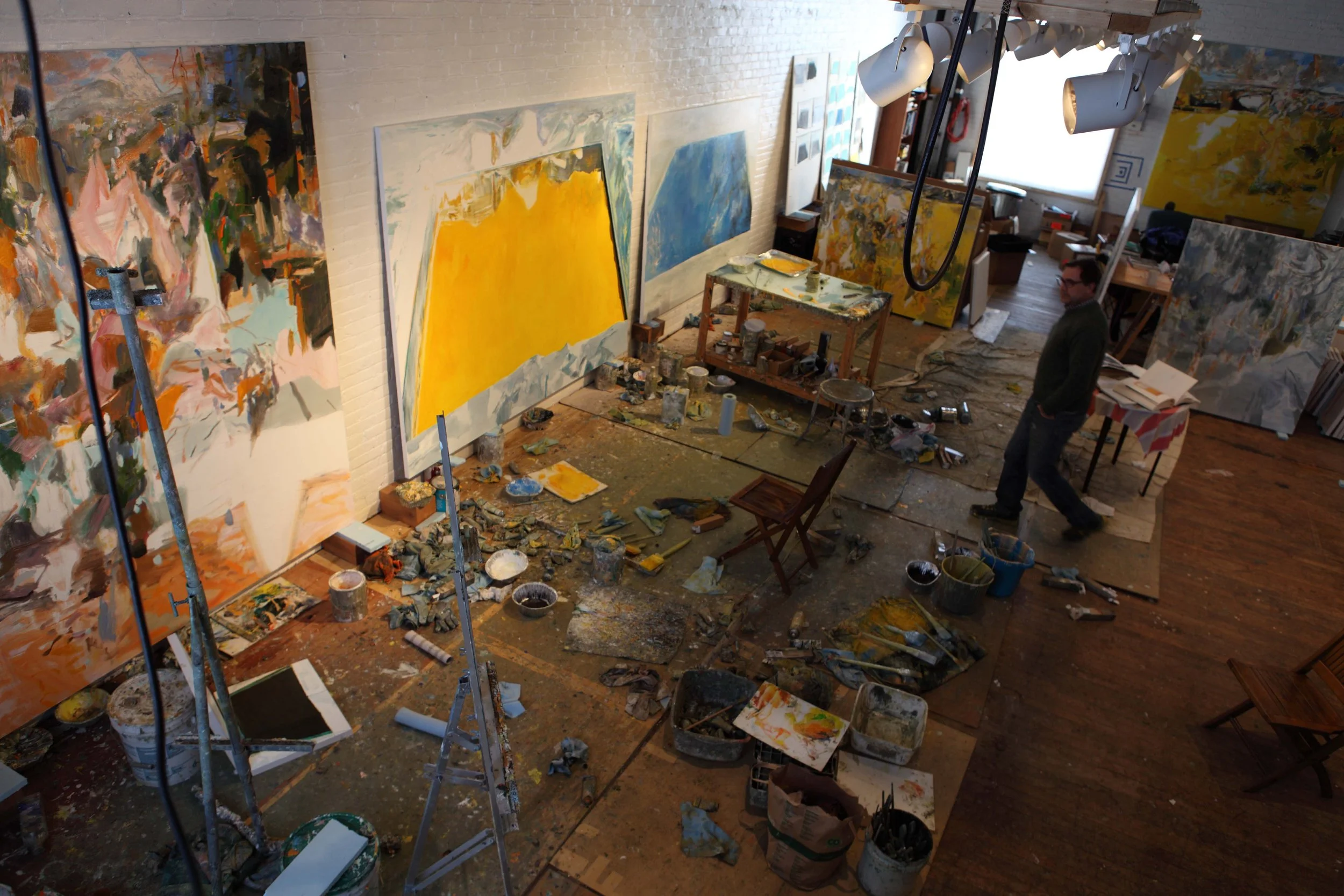 An art studio filled with large abstract paintings, scattered painting supplies, and a person walking through the cluttered space.