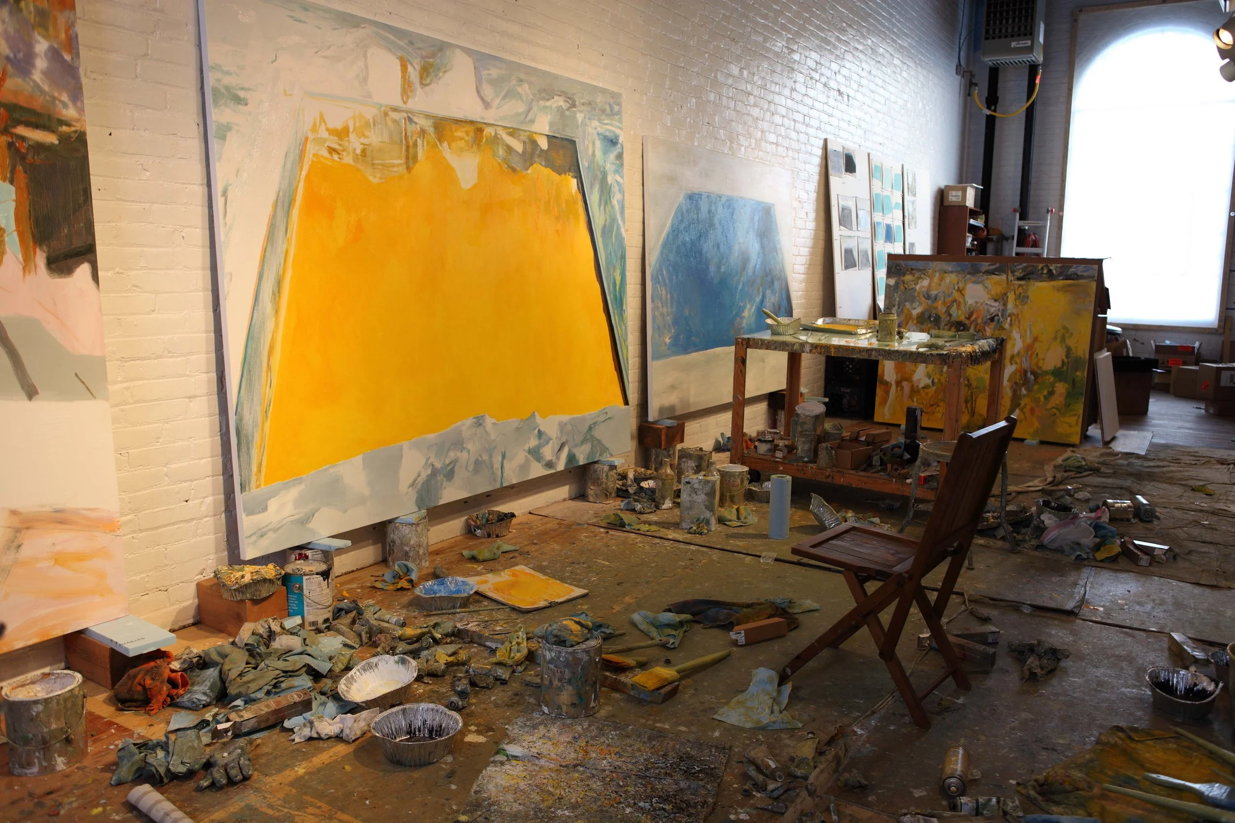 An art studio with large abstract paintings on the white brick walls, an art table with paints and brushes, and scattered painting supplies on the floor.
