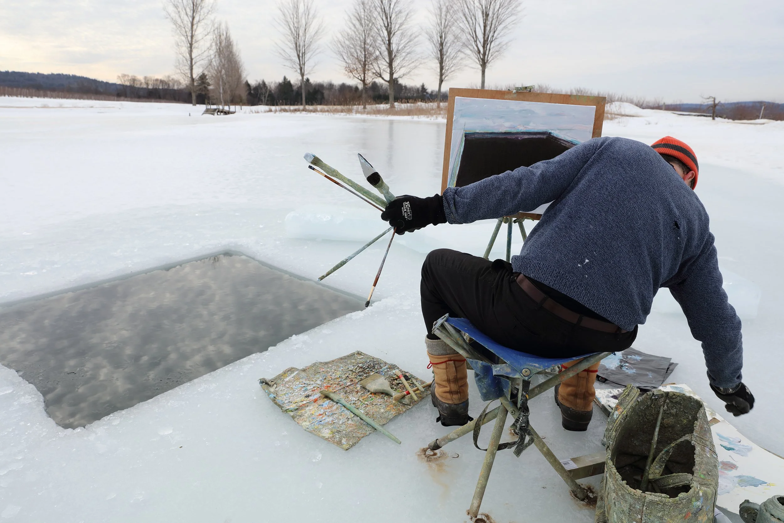 A person in winter clothing is shaping a hole in the ice of a frozen lake, with an easel and painting supplies nearby, in a snowy outdoor setting.