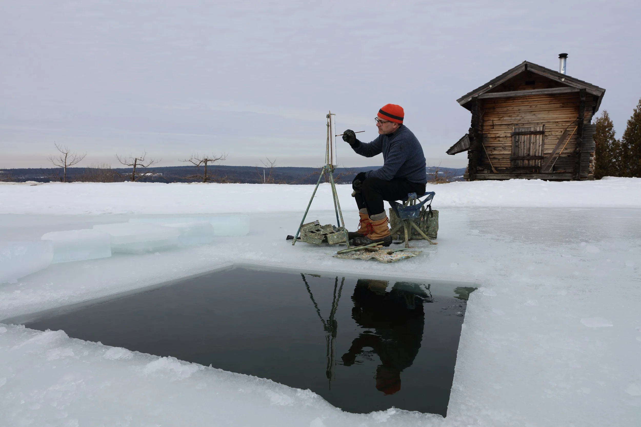 A man in winter clothing, including an orange beanie, glasses, and gloves, is sitting on a small chair on a frozen lake, fishing through a hole in the ice. There is a small wooden house and leafless trees in the background under a cloudy sky.