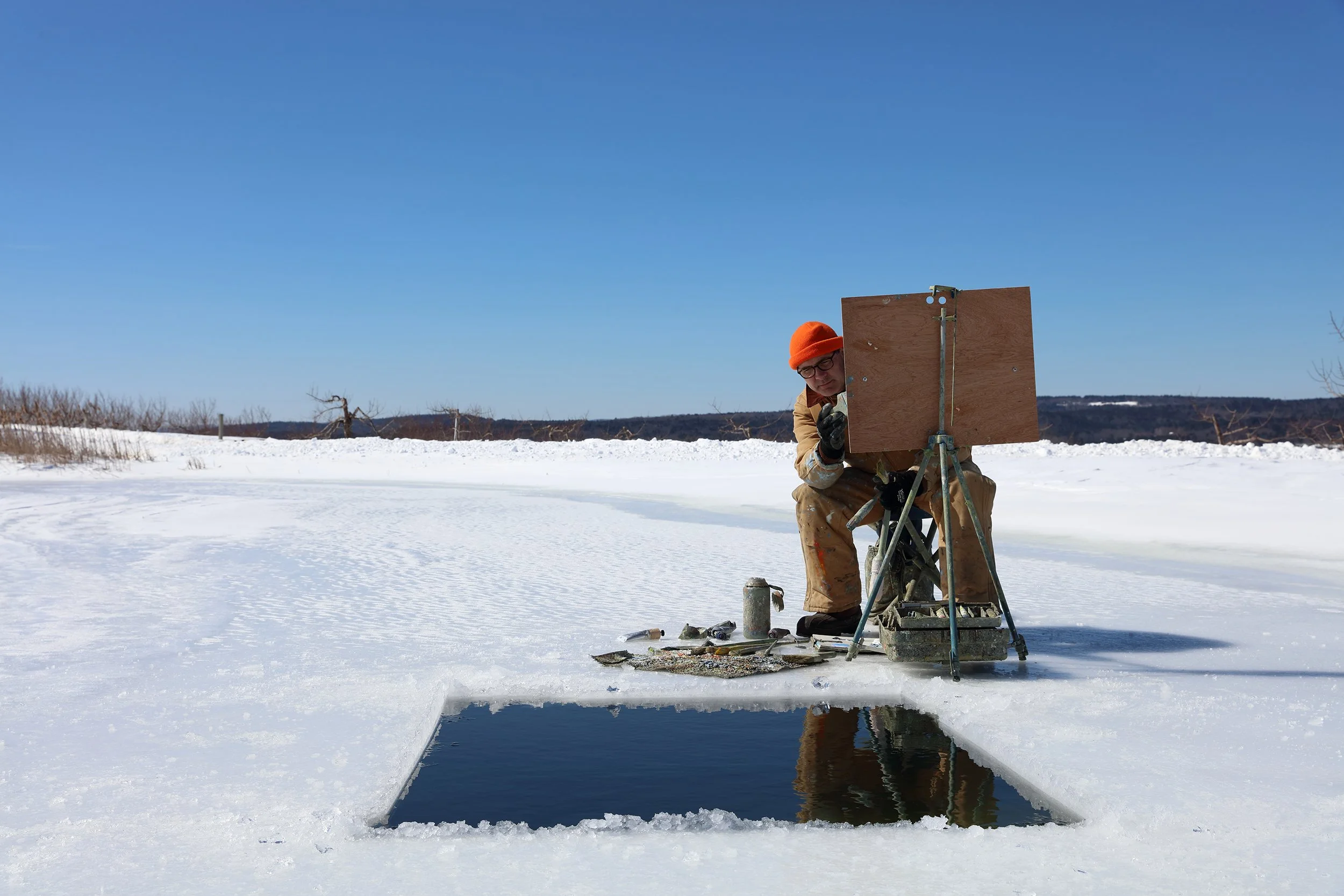 A person wearing an orange beanie, glasses, and winter clothing setting up equipment on snow-covered ground with a clear blue sky and distant trees in the background.