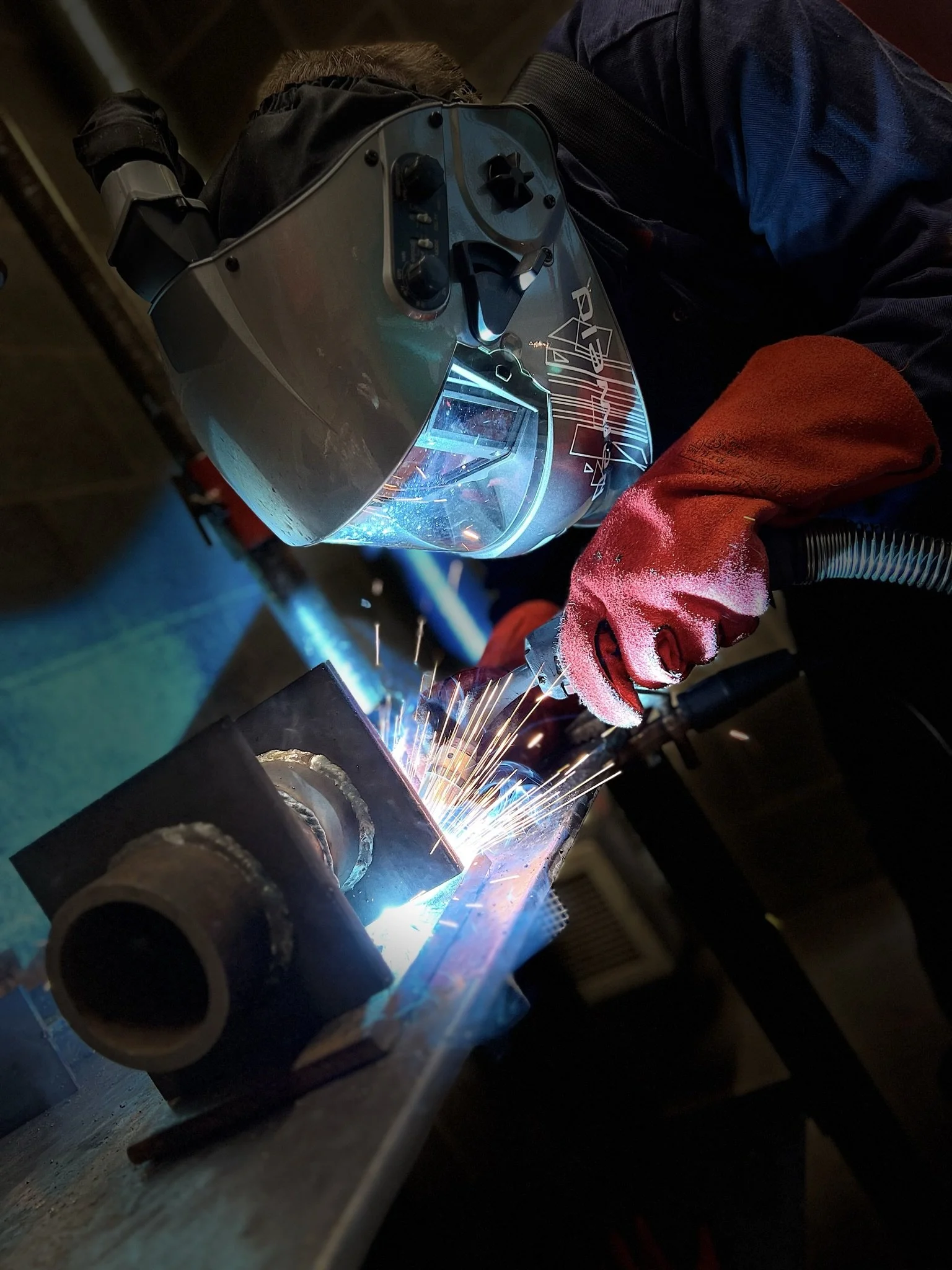 A person welding metal pipe with bright sparks and blue welding light, wearing a protective helmet and red glove.