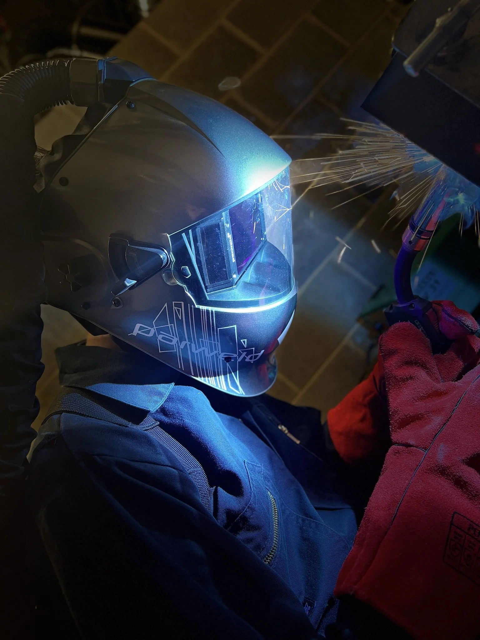 A person wearing a protective welding helmet with a blue tint, also wearing a dark jacket with a white and blue design, is welding metal. Sparks are flying from the welding process, and a gloved hand is holding a tool while working on the metal.