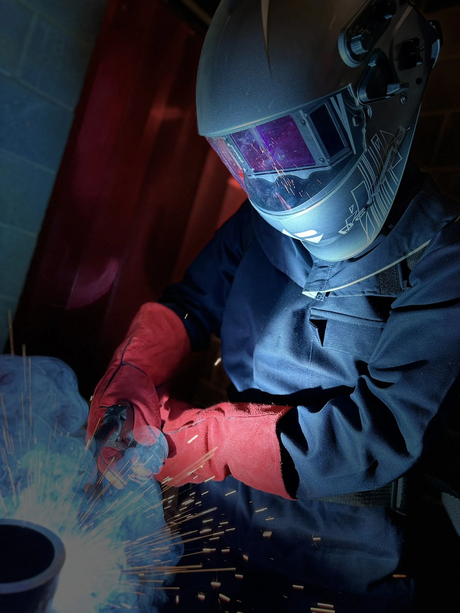 Person wearing a welding helmet working on a metal project, sparks flying in the air, indoors with a blue wall in the background.