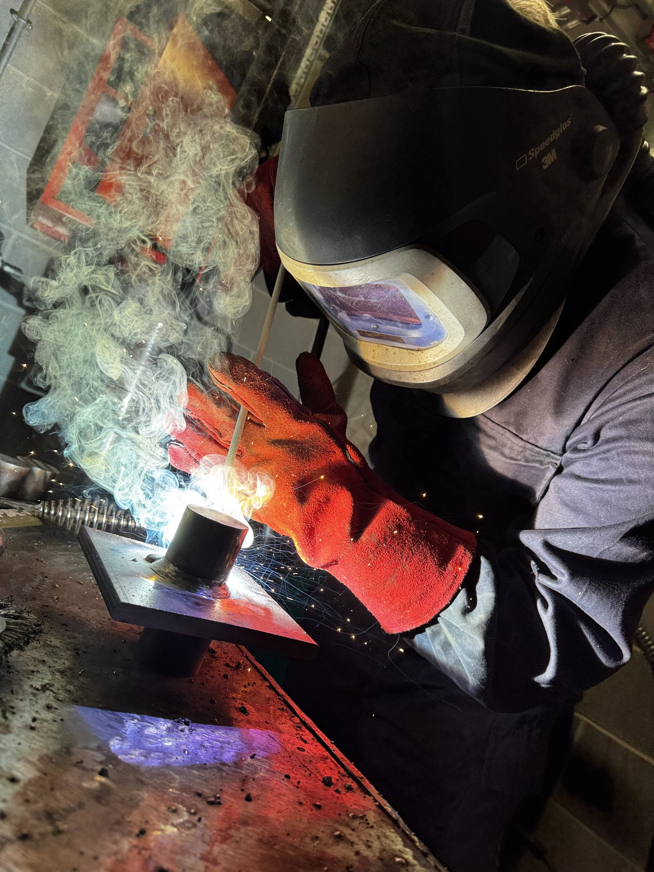 A person wearing a welding helmet and gloves welding a metal piece, with sparks and smoke.