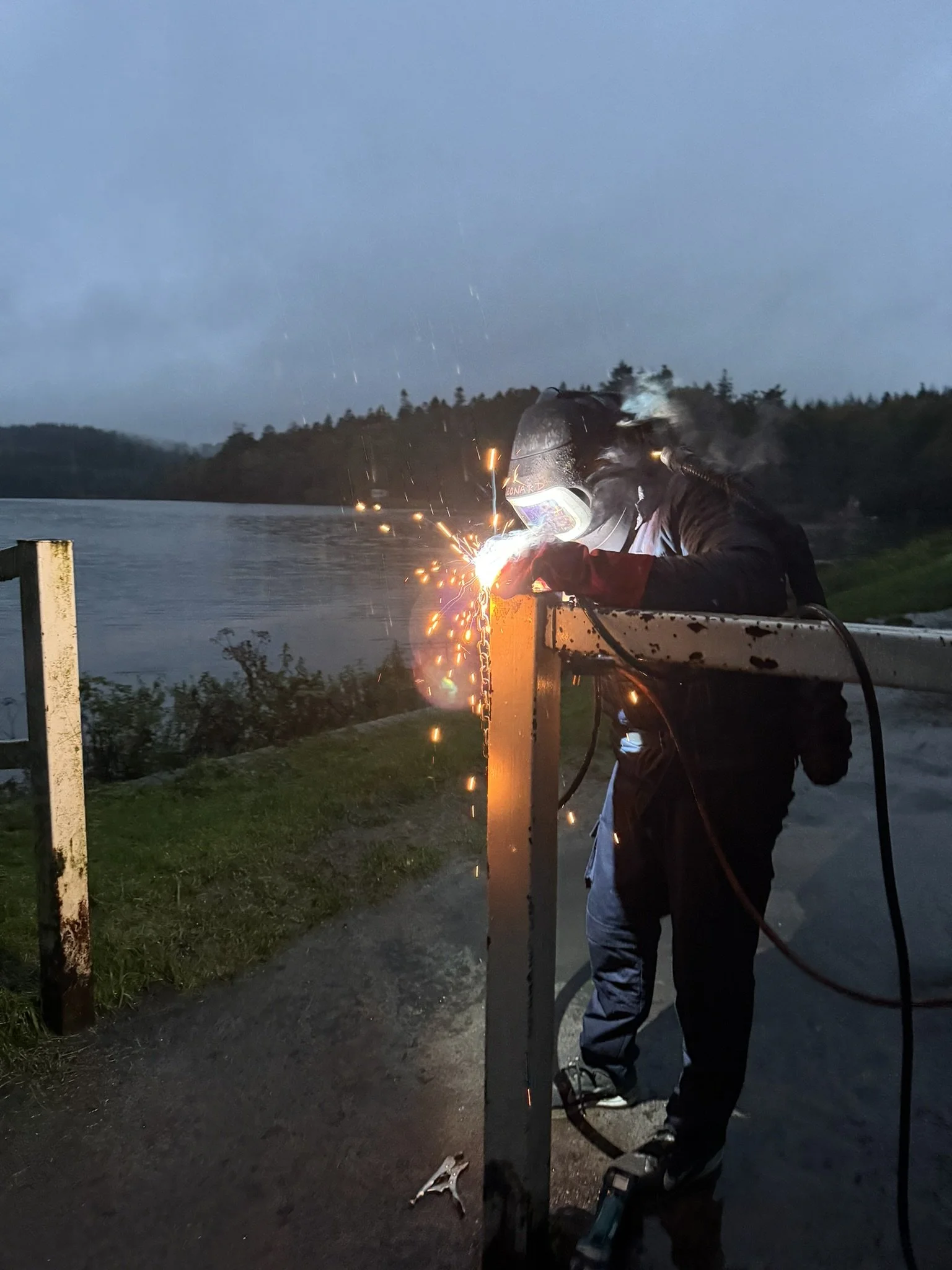 A person welding metal post by a body of water during the evening, with sparks flying and wearing protective gear.