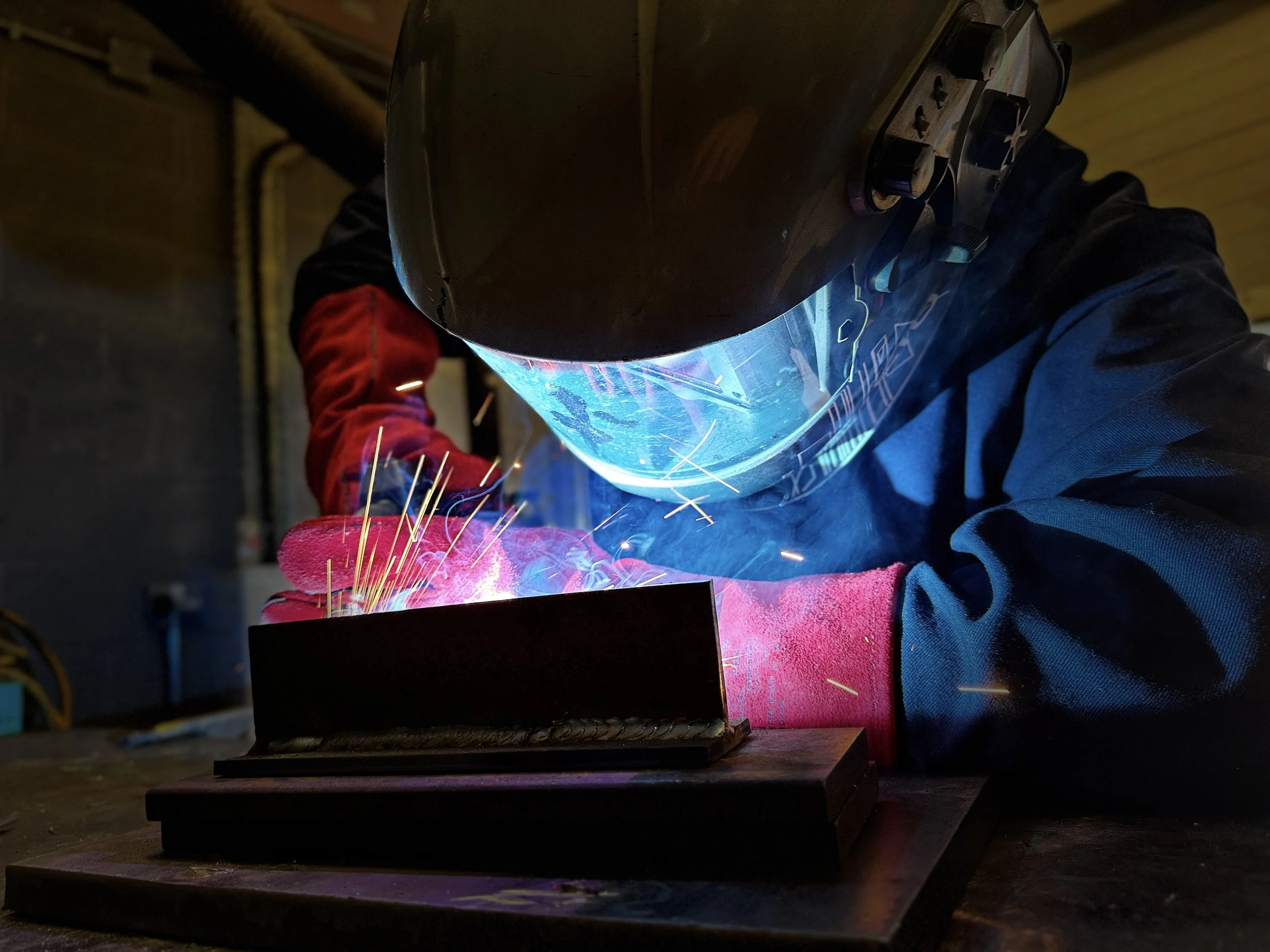 A person welding metal with a protective helmet and gloves, sparks flying from the welding process, in an industrial setting.
