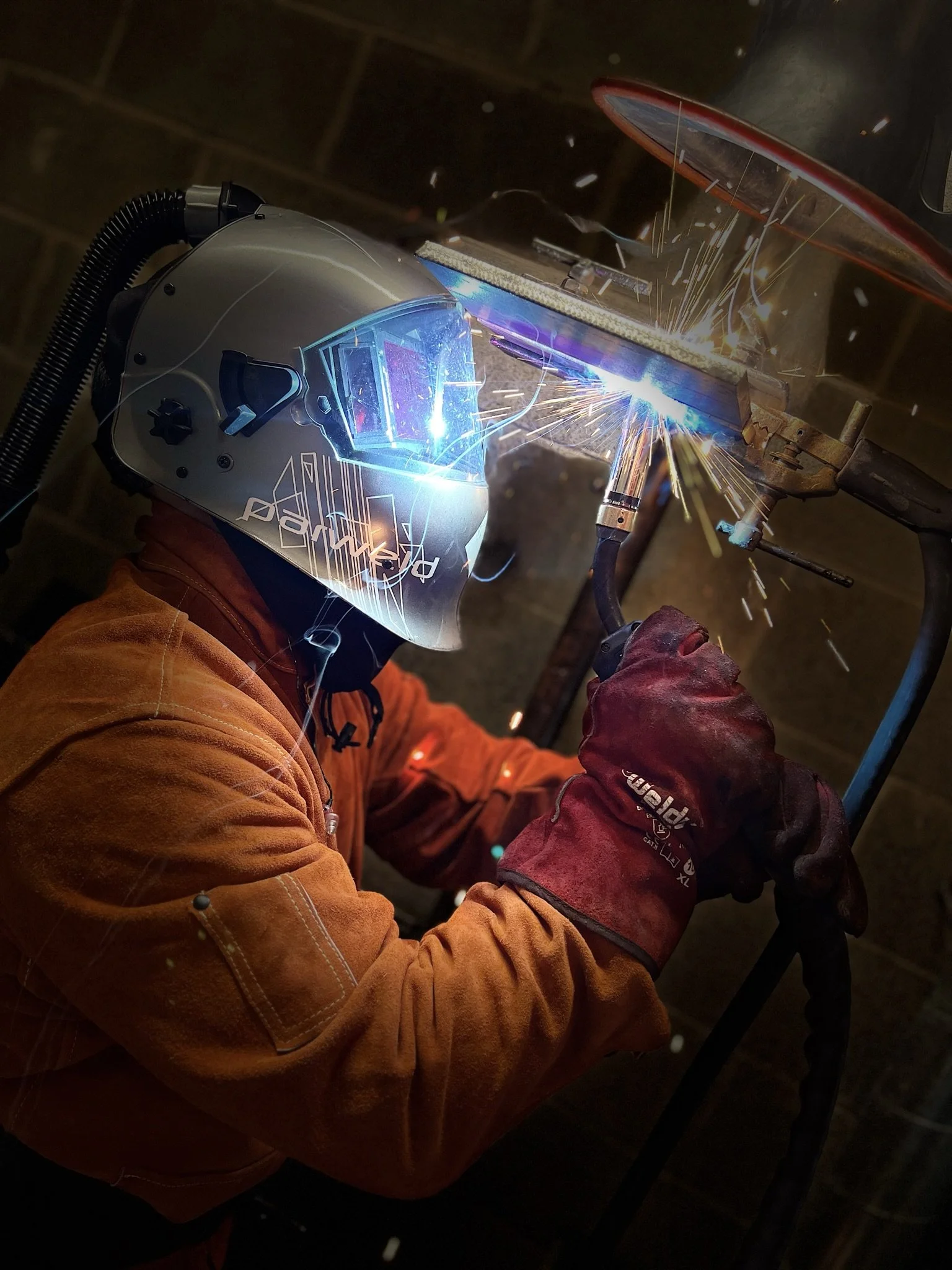 Worker welding metal with sparks flying near their helmet and protective gloves in a workshop or construction environment.