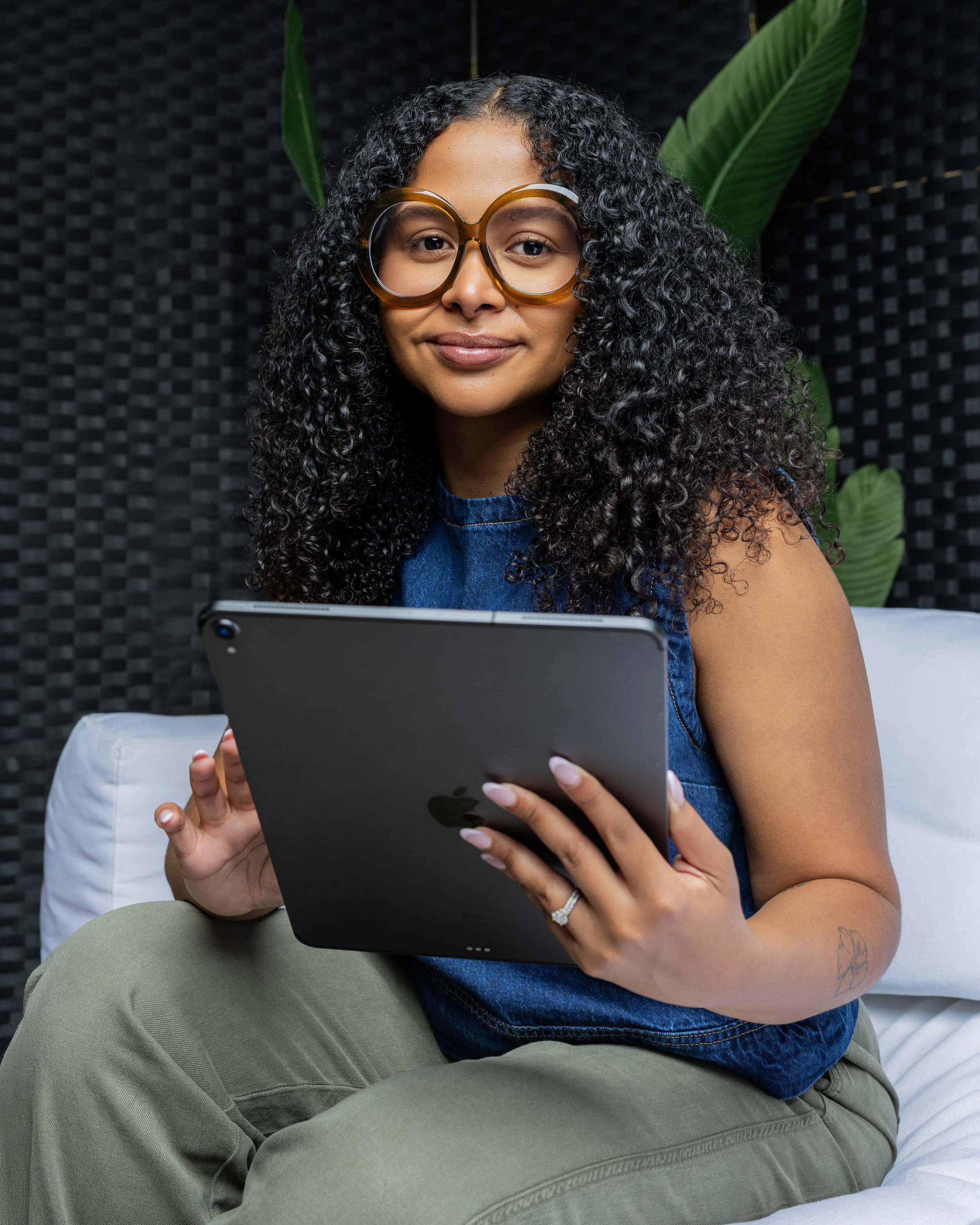 A woman with curly black hair, wearing large glasses and a denim top, sitting with her legs crossed, smiling while holding a tablet. She has a tattoo of a butterfly on her left arm and is sitting in a room with a black textured wall and large green plants in the background.