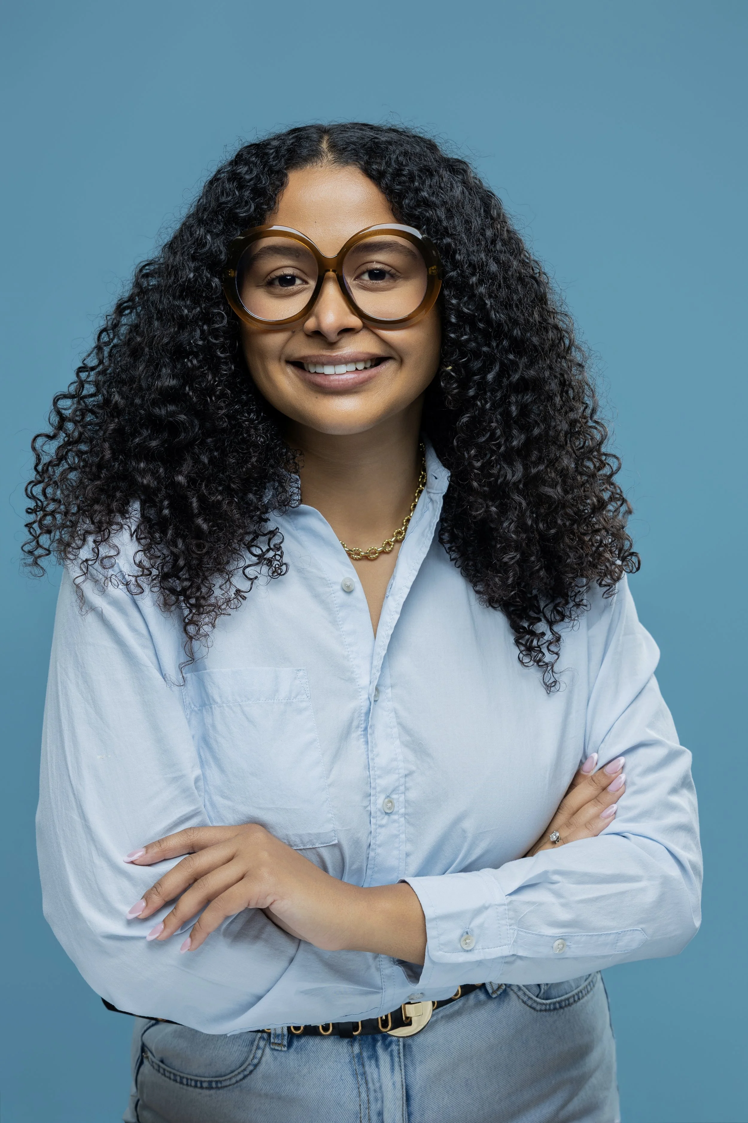 A woman with curly black hair, wearing large glasses, a light blue button-down shirt, a gold chain necklace, and jeans, standing against a blue background with her arms crossed and smiling.