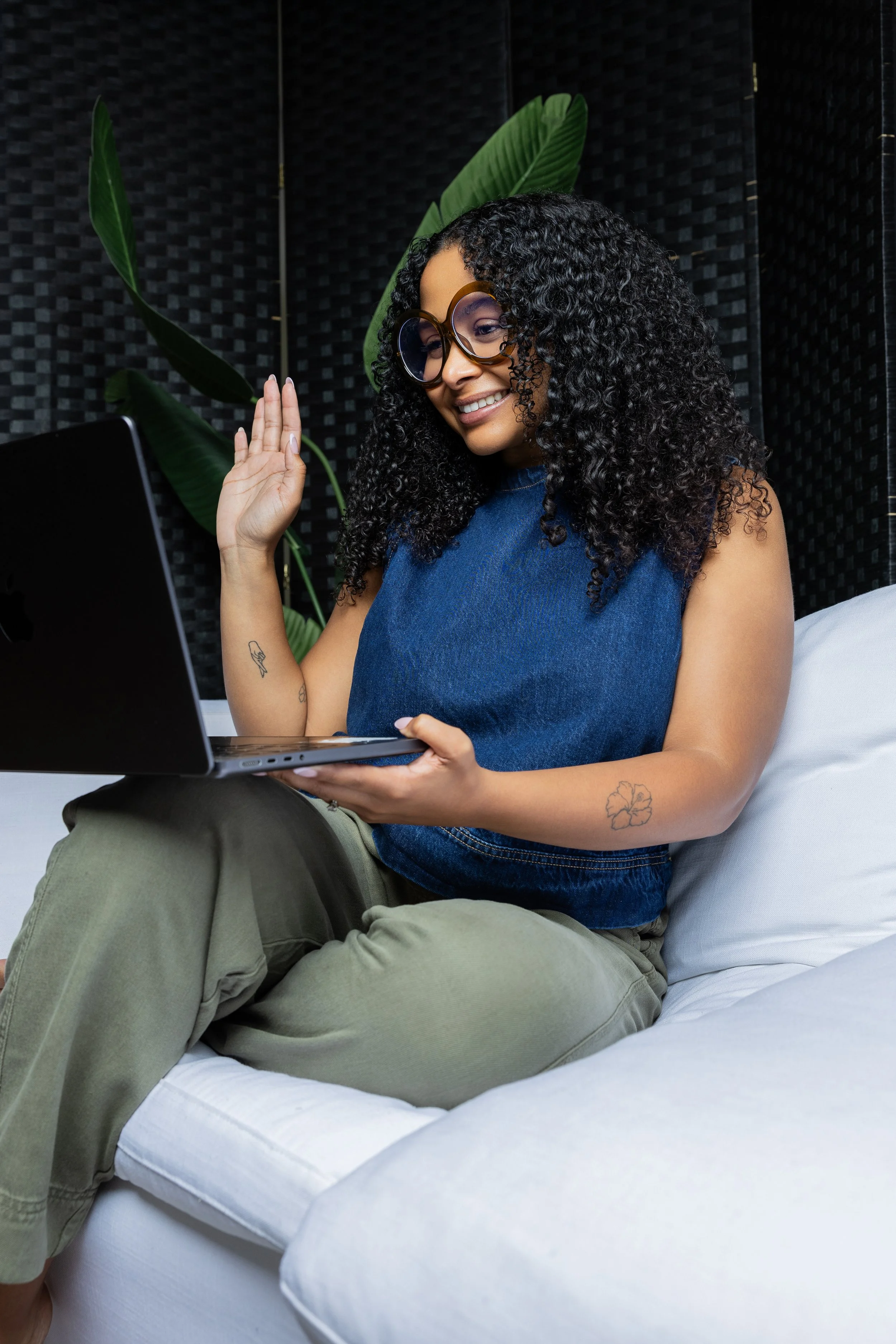 A woman sitting on a bed, smiling and waving at a laptop screen, wearing large sunglasses and casual clothes, with large green tropical leaves in the background.