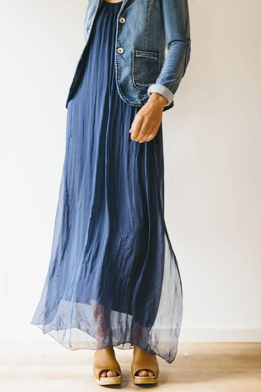 A person wearing a long navy blue pleated skirt, a blue denim jacket with rolled cuffs, and tan platform sandals, standing indoors against a plain white wall.
