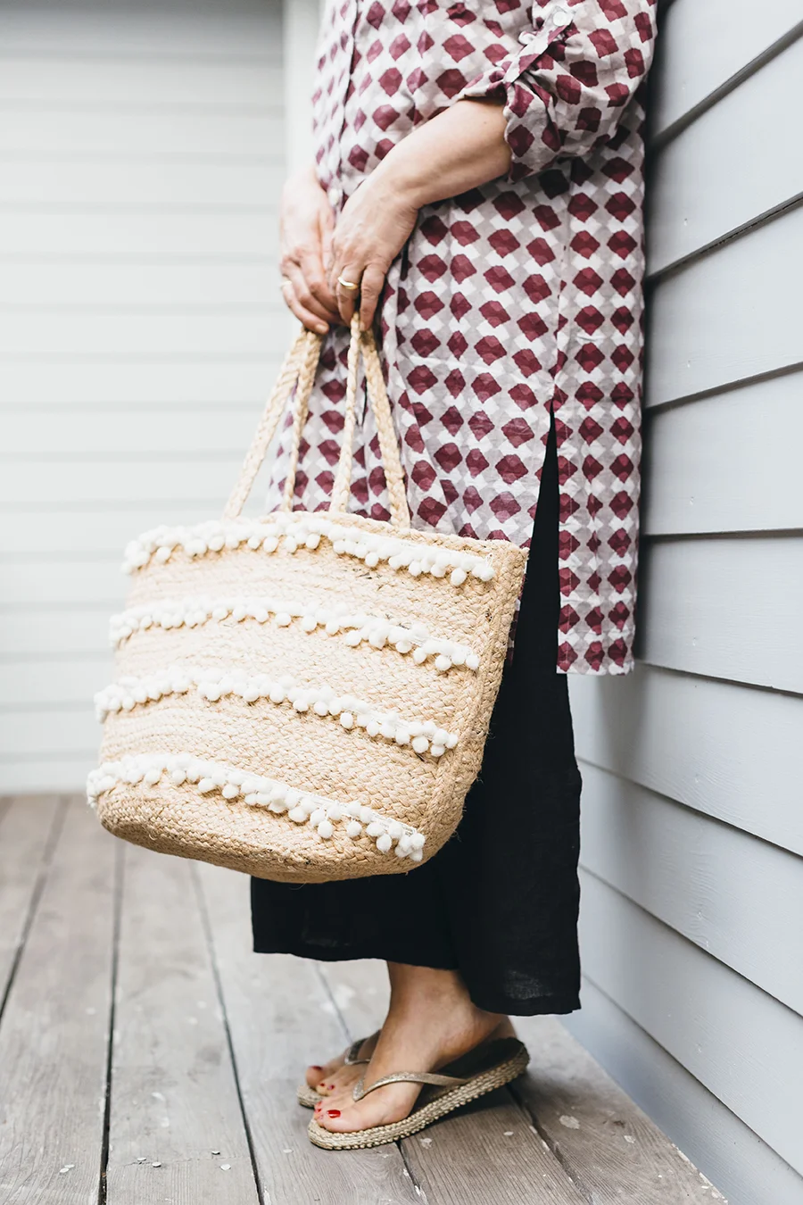 A person wearing black pants and flip-flops holding a woven straw tote bag with white pom-pom accents. They are standing on a wooden deck next to a gray siding wall, and only part of their body is visible.