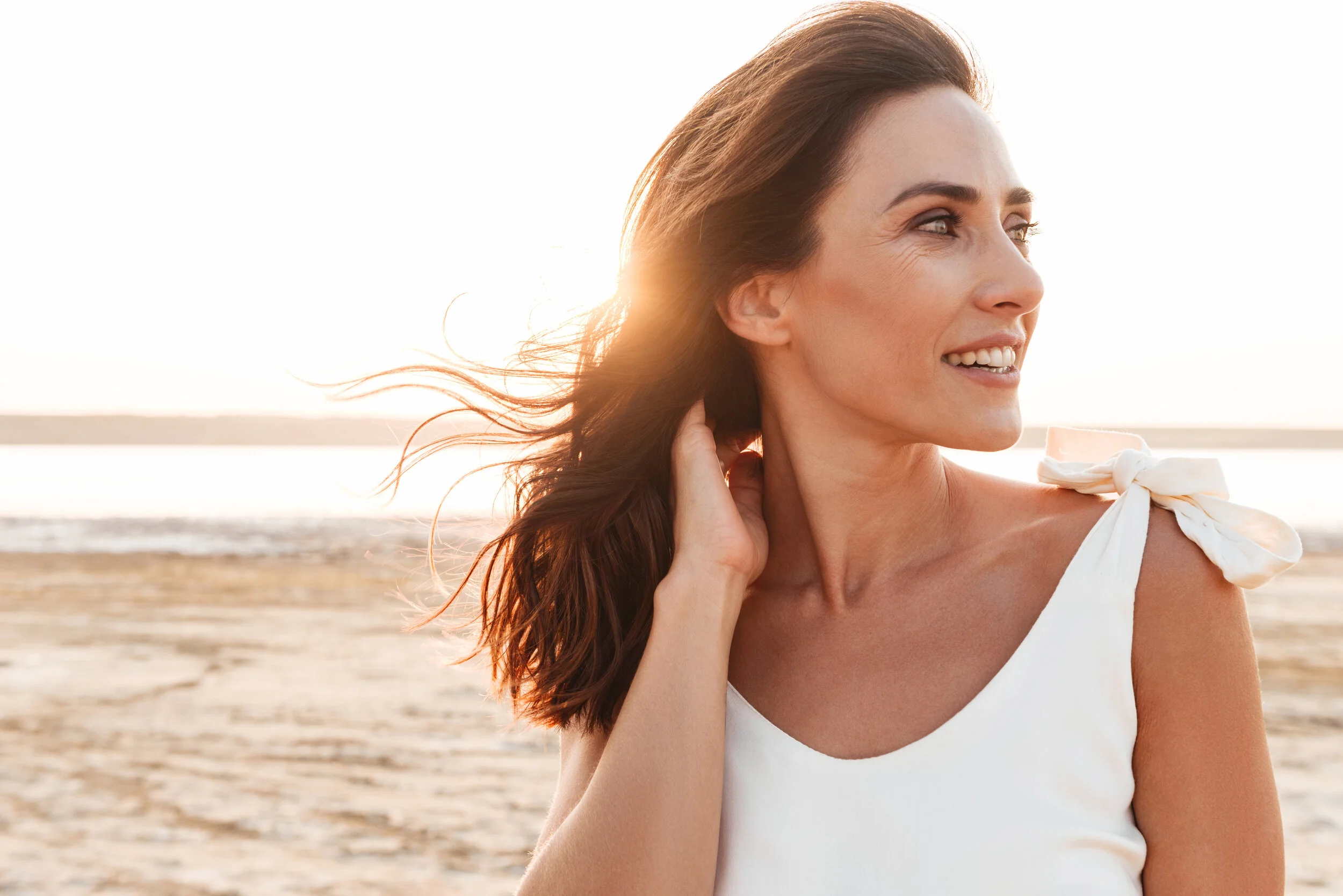 A woman with long brown hair smiling and looking to her right while standing on a beach at sunset.