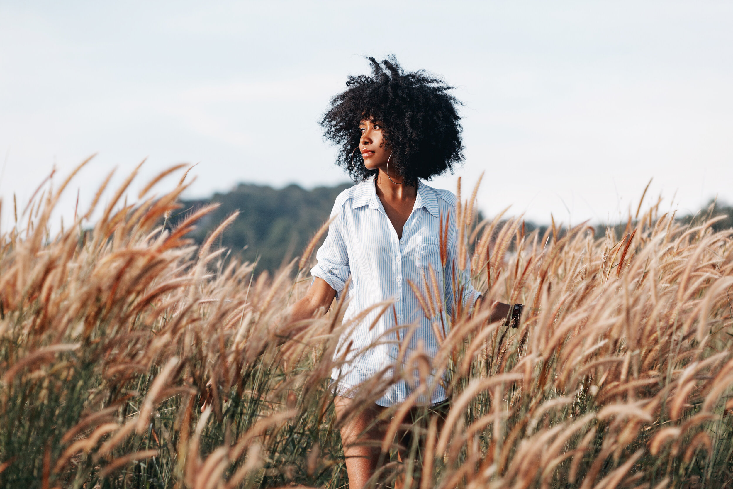 A woman with curly hair wearing a striped shirt standing in a field of tall grass, looking to the side.