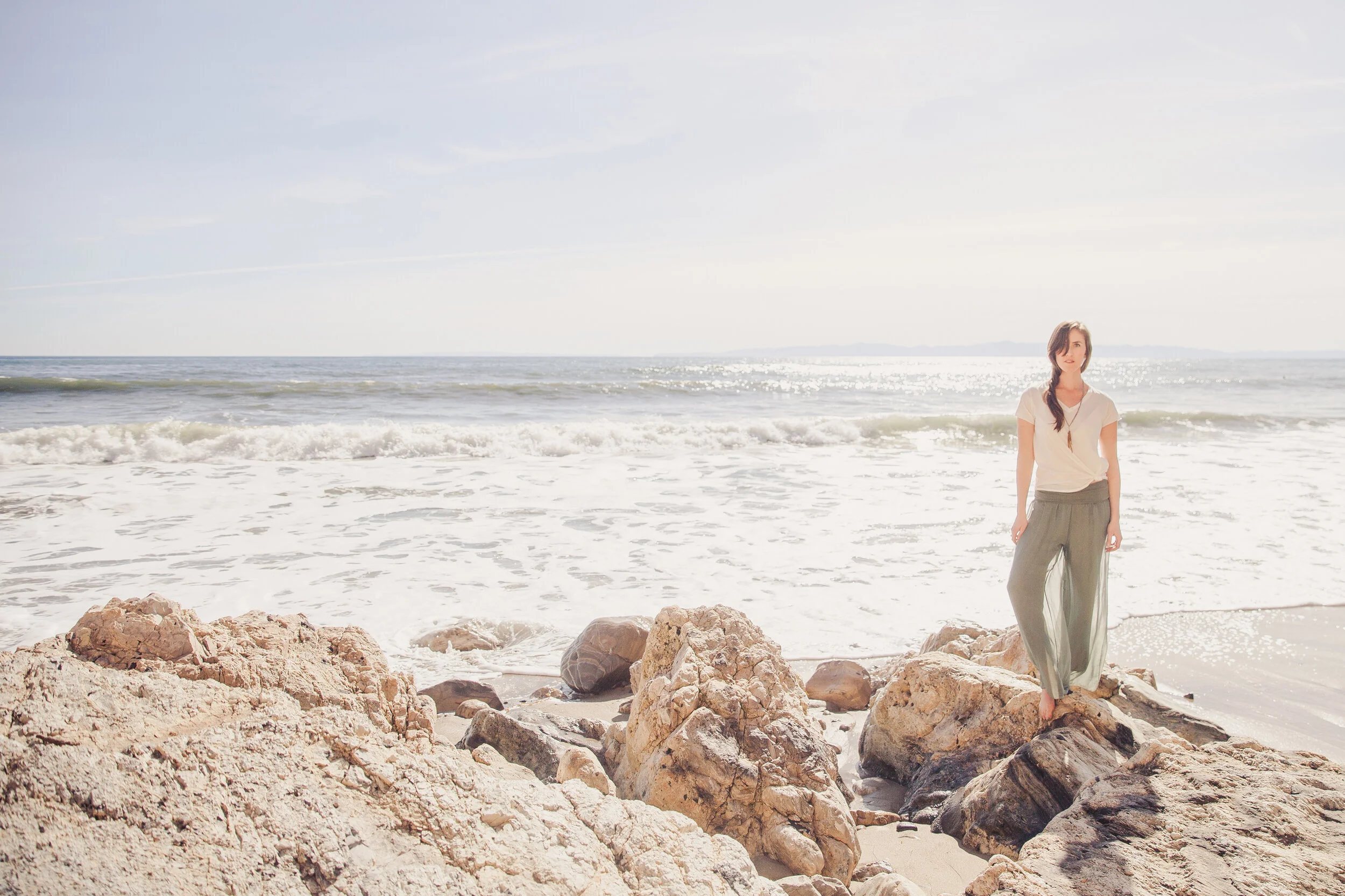 A woman standing on rocks at the beach near the ocean, wearing a white top and flowy pants, with the sun shining and waves in the background.