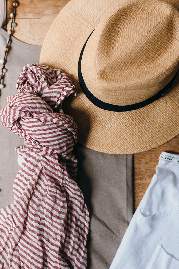 A beige straw hat with a black band, a red and white striped cloth, a white shirt, and part of a necklace are laid out on a wooden surface.