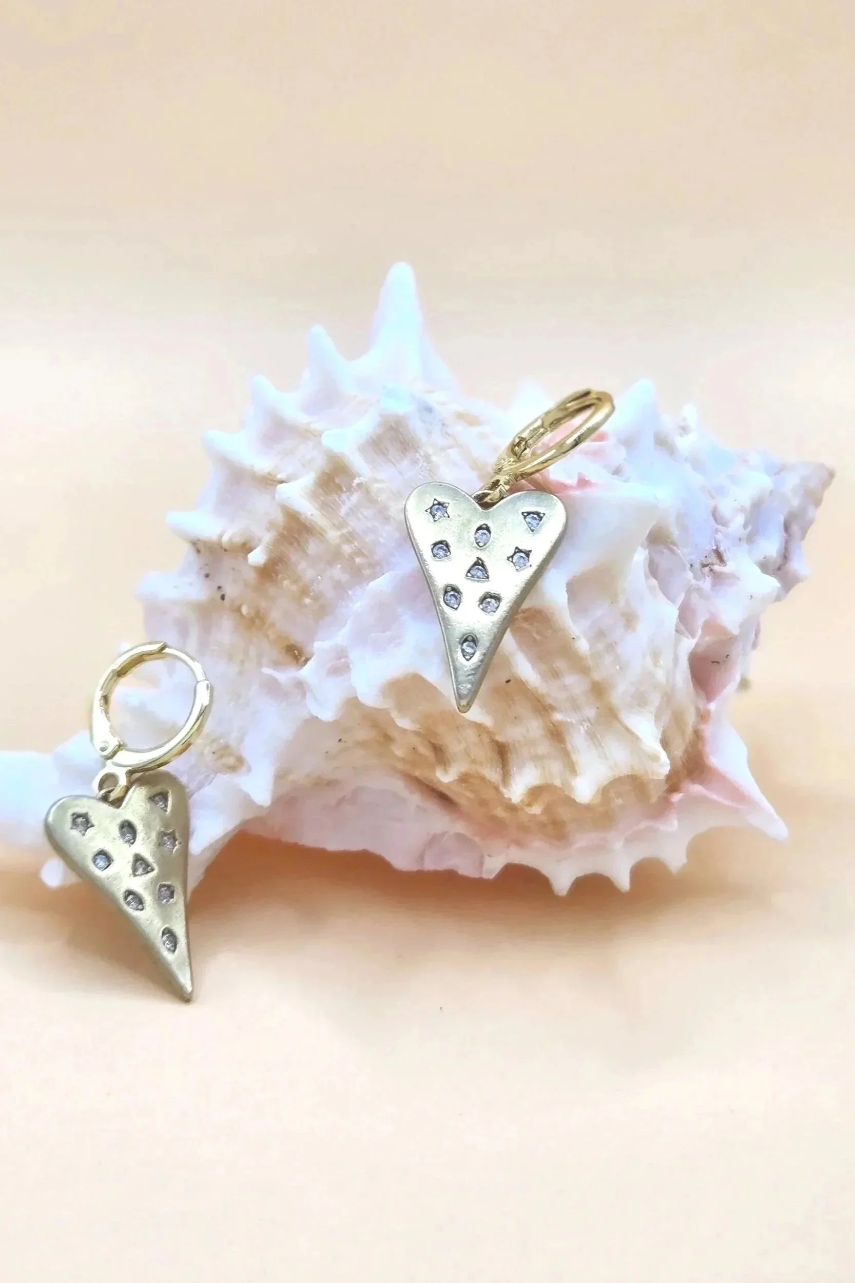 Close-up of a white seashell with a small, gold heart-shaped pendant hanging on it. The pendant is decorated with small, star-shaped cutouts filled with blue stones. A second similar pendant, partially visible, hangs in the foreground. The background is plain and light-colored.
