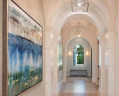 Elegant hallway with arched ceiling, white walls, and large paintings, featuring a window at the end with a view of greenery.