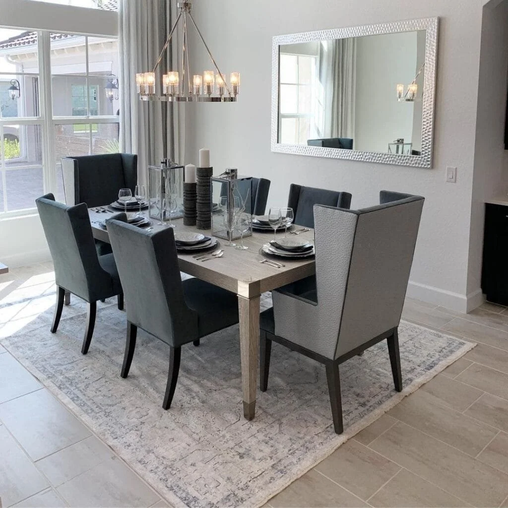 Modern dining room with a rectangular table set with black and white dinnerware, glassware, and decorated with tall candles. The room features a large mirror on the wall, a chandelier overhead, and large windows with curtains allowing natural light.