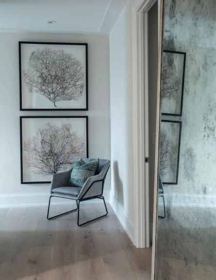 A modern interior corner with two framed black-and-white tree prints on the wall, a gray armchair with two patterned pillows, and a mirror reflecting part of the wall and similar art.